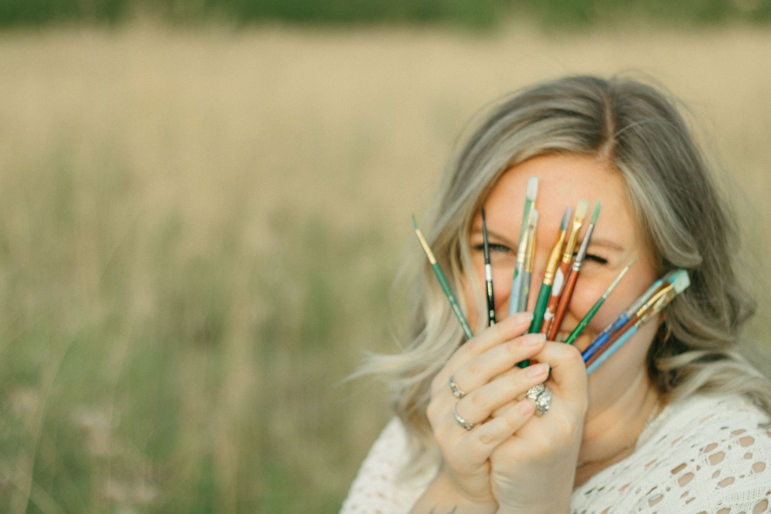 Smiling woman playfully hiding behind a fan of colorful paintbrushes in a meadow.