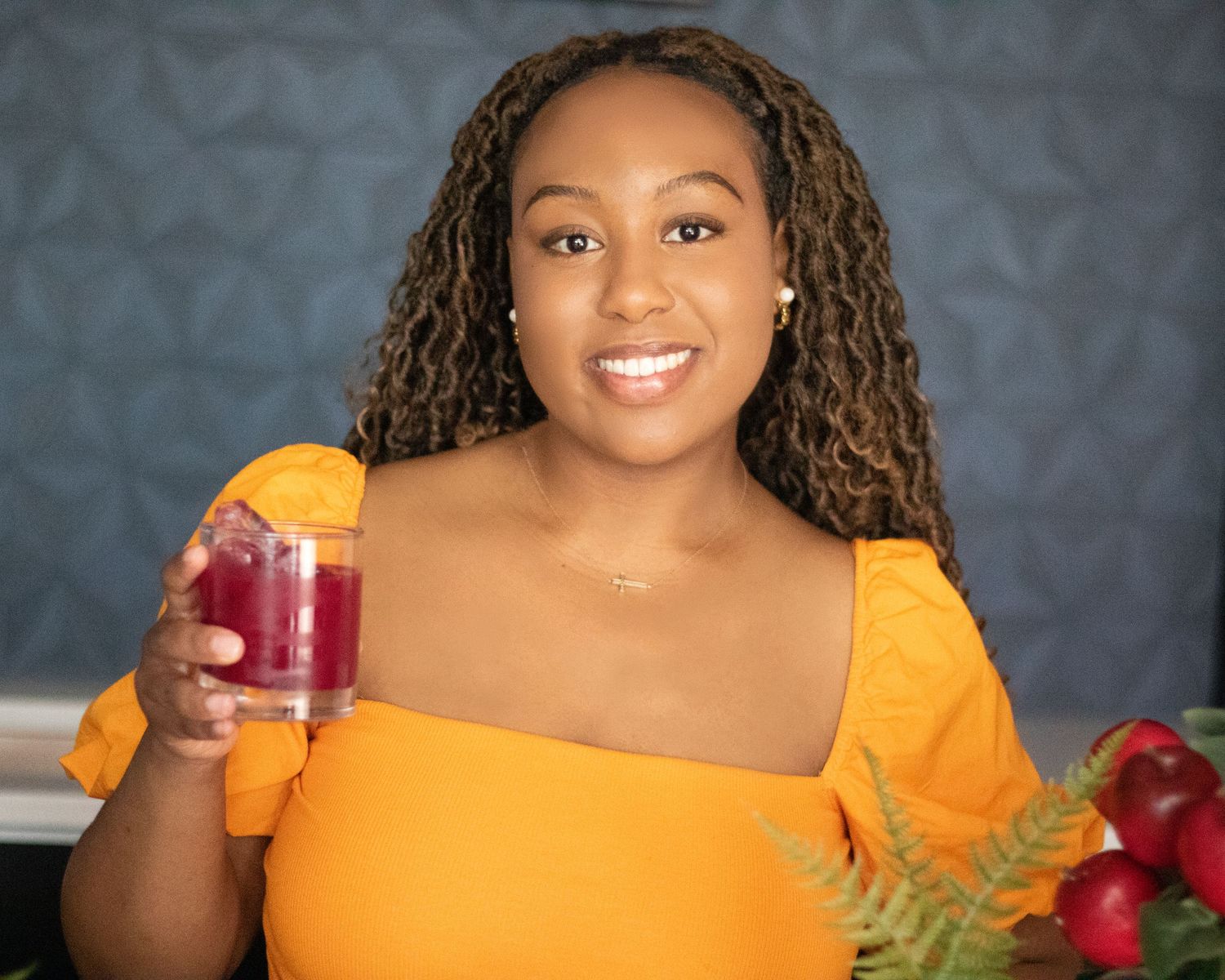 Smiling woman in yellow off-shoulder top holds a glass of red juice surrounded by fresh fruits.