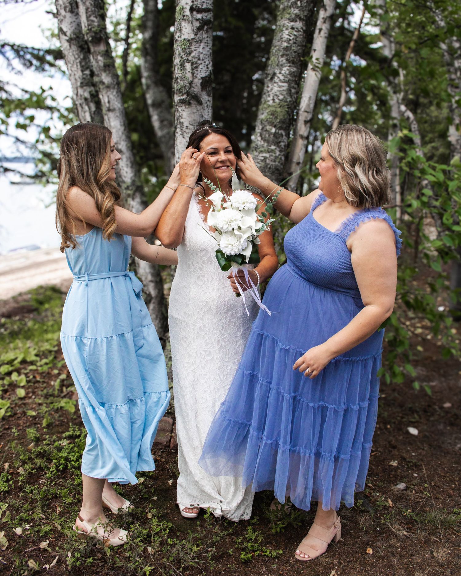 Bride in white lace dress holding bouquet laughs with two bridesmaids in blue dresses among trees.