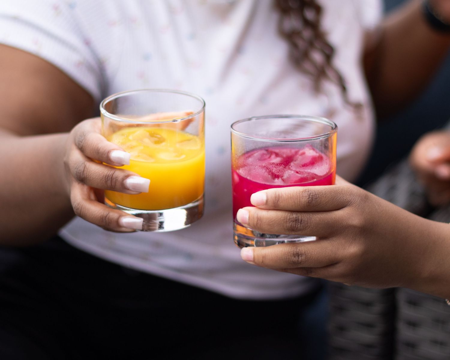 Two hands clinking glasses of vibrant orange and pink iced juice drinks.