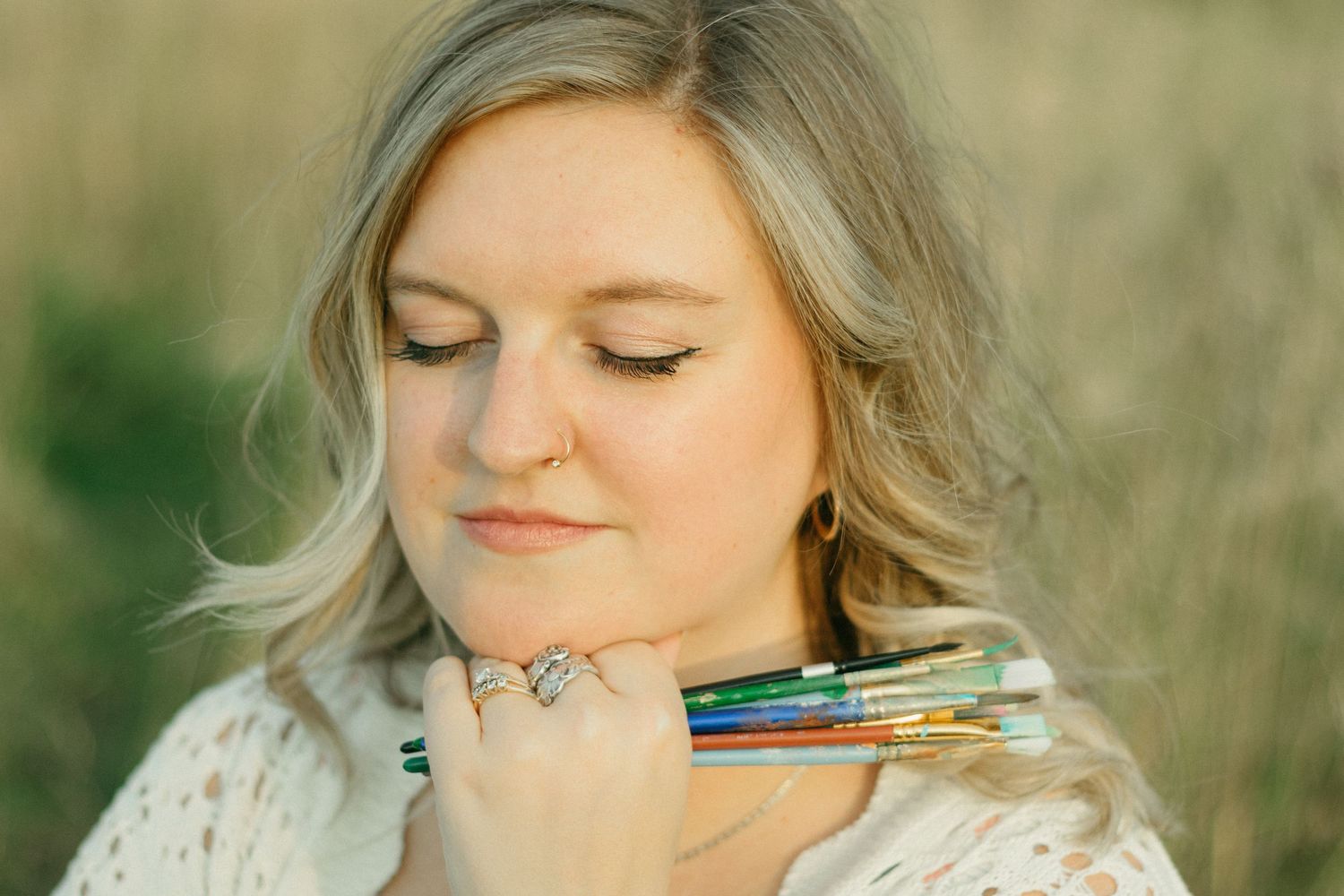Blonde artist smiling softly while holding colorful paintbrushes against her chin outdoors.