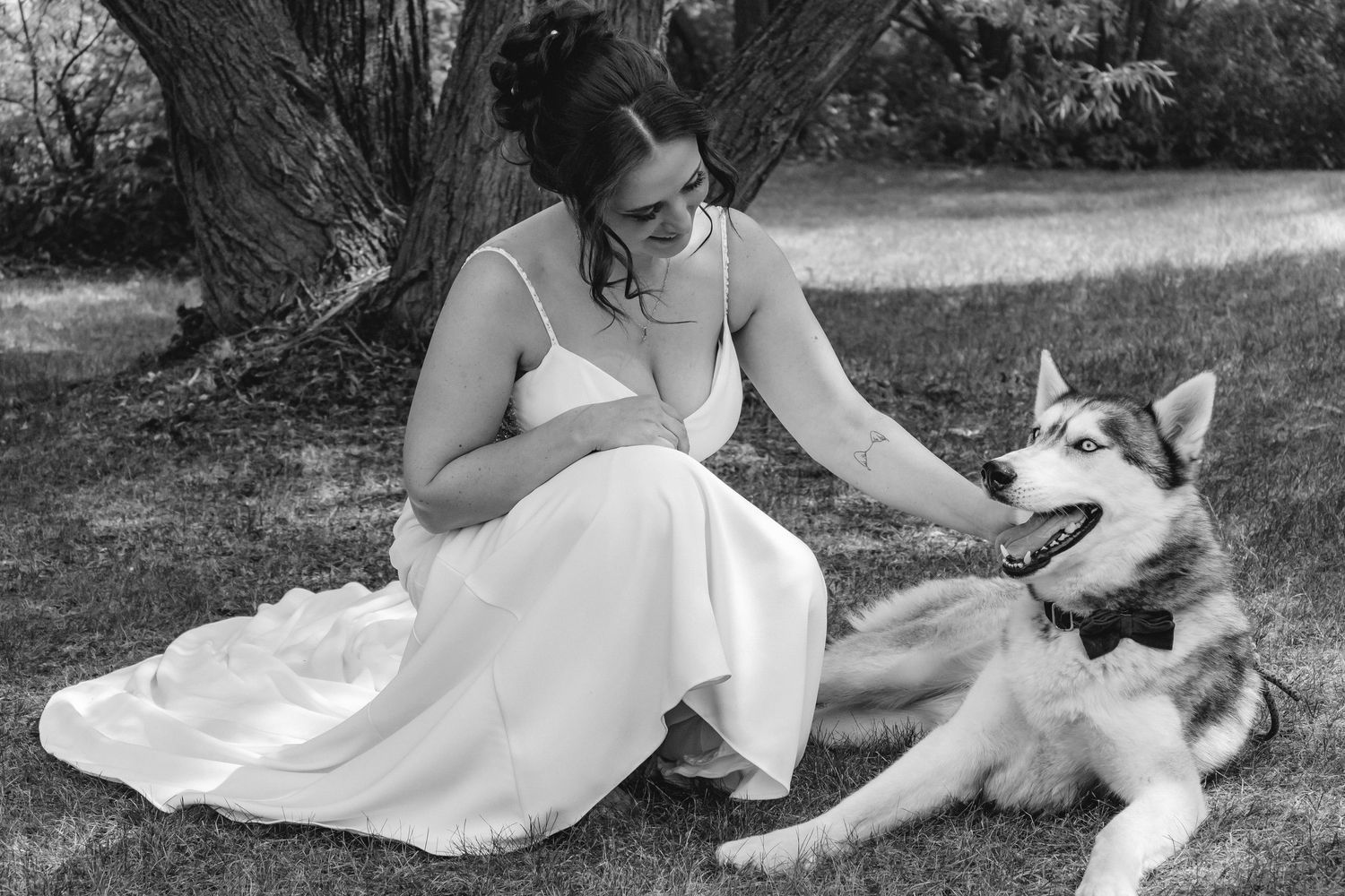 Black and white photo of bride in white gown sitting on grass petting a husky dog.