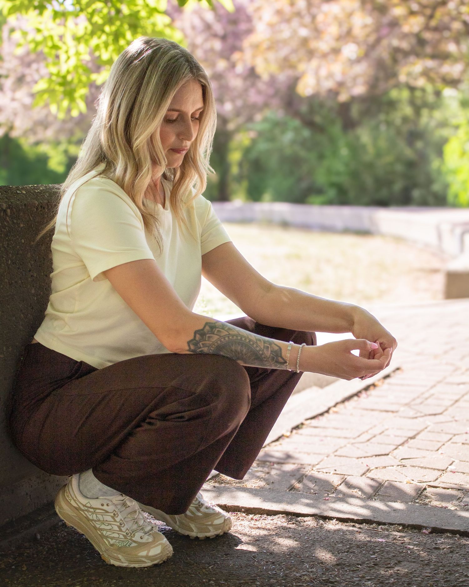 Blonde woman with tattoos crouching outdoors in white tee and brown pants near blooming trees.