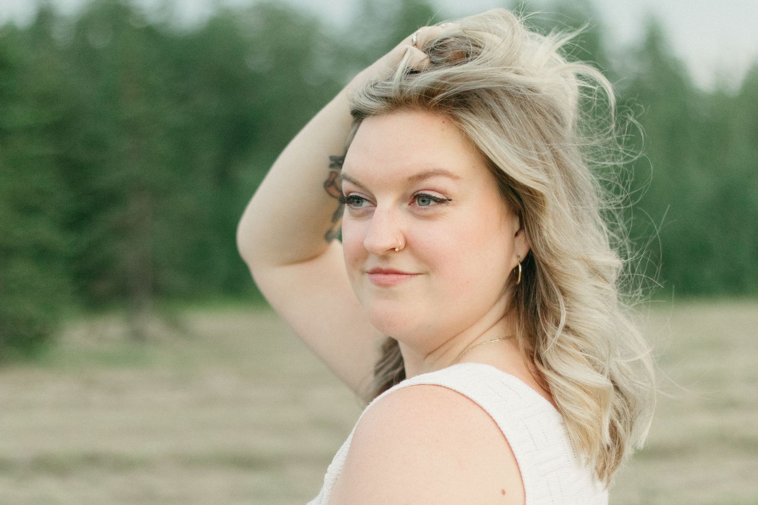 Blonde woman in white tank top smiling over her shoulder in a green meadow.
