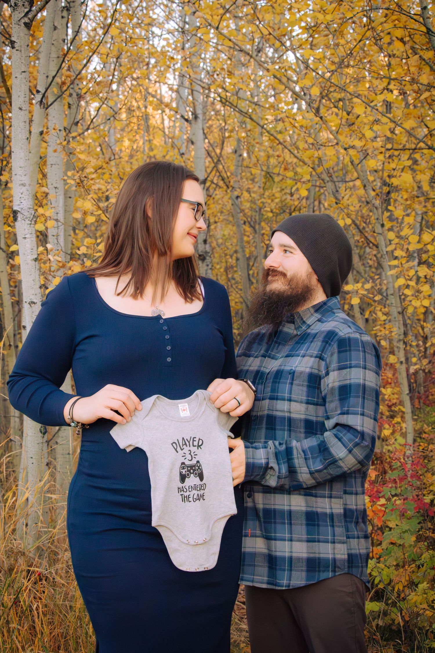 Pregnant couple holding baby onesie with gaming print among colorful fall foliage.
