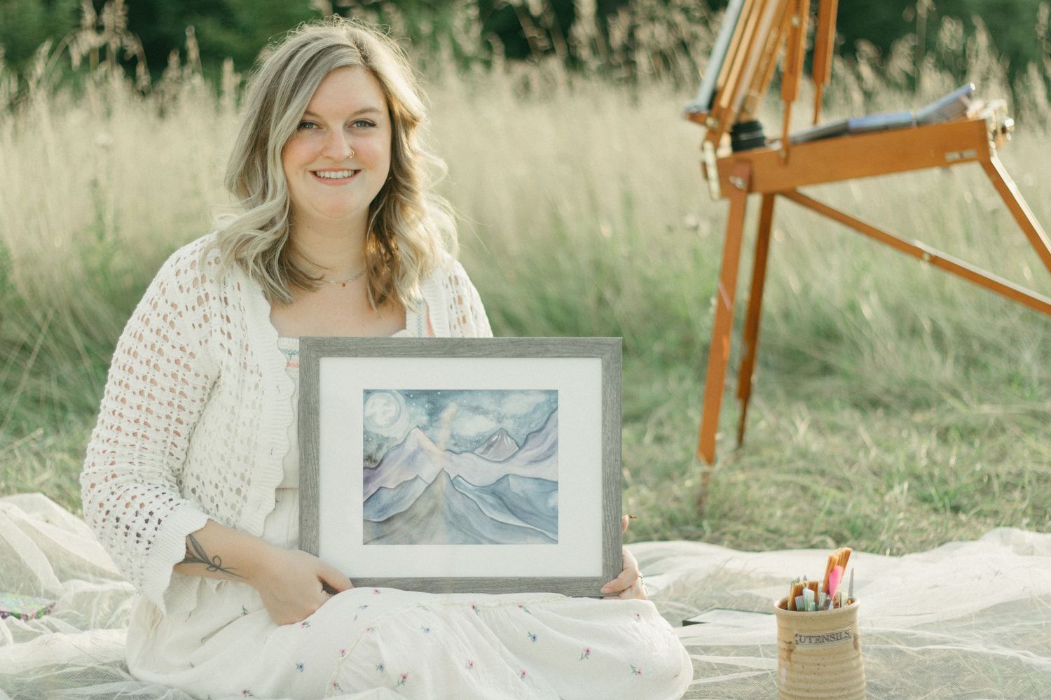 Smiling blonde woman sitting outdoors holding framed watercolor painting on blanket.