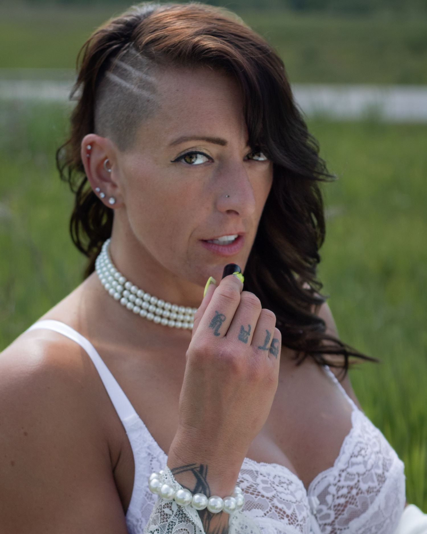 A woman with a shaved side hairstyle wearing pearl jewelry and white lace poses outdoors in a field.