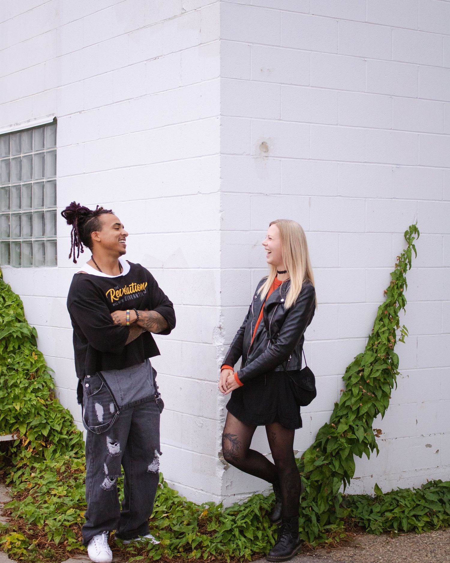 Two young people laughing and conversing against a white brick wall with ivy, one in a black hoodie and one in a leather jacket.