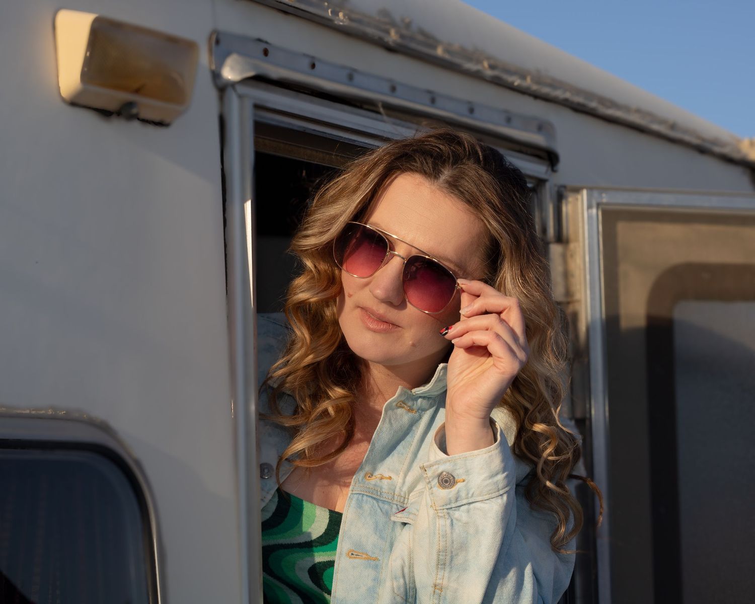 Woman with curly hair and pink aviator sunglasses leaning out of a vintage camper trailer window.