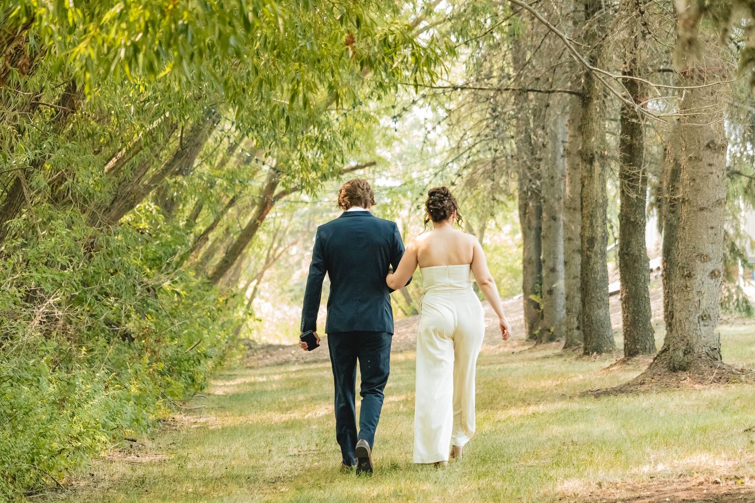 Bride and groom walking hand in hand along a tree-lined path on their wedding day.