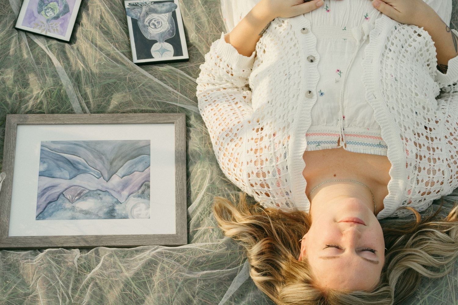 Overhead view of blonde woman in knit sweater lying beside framed artwork outdoors.