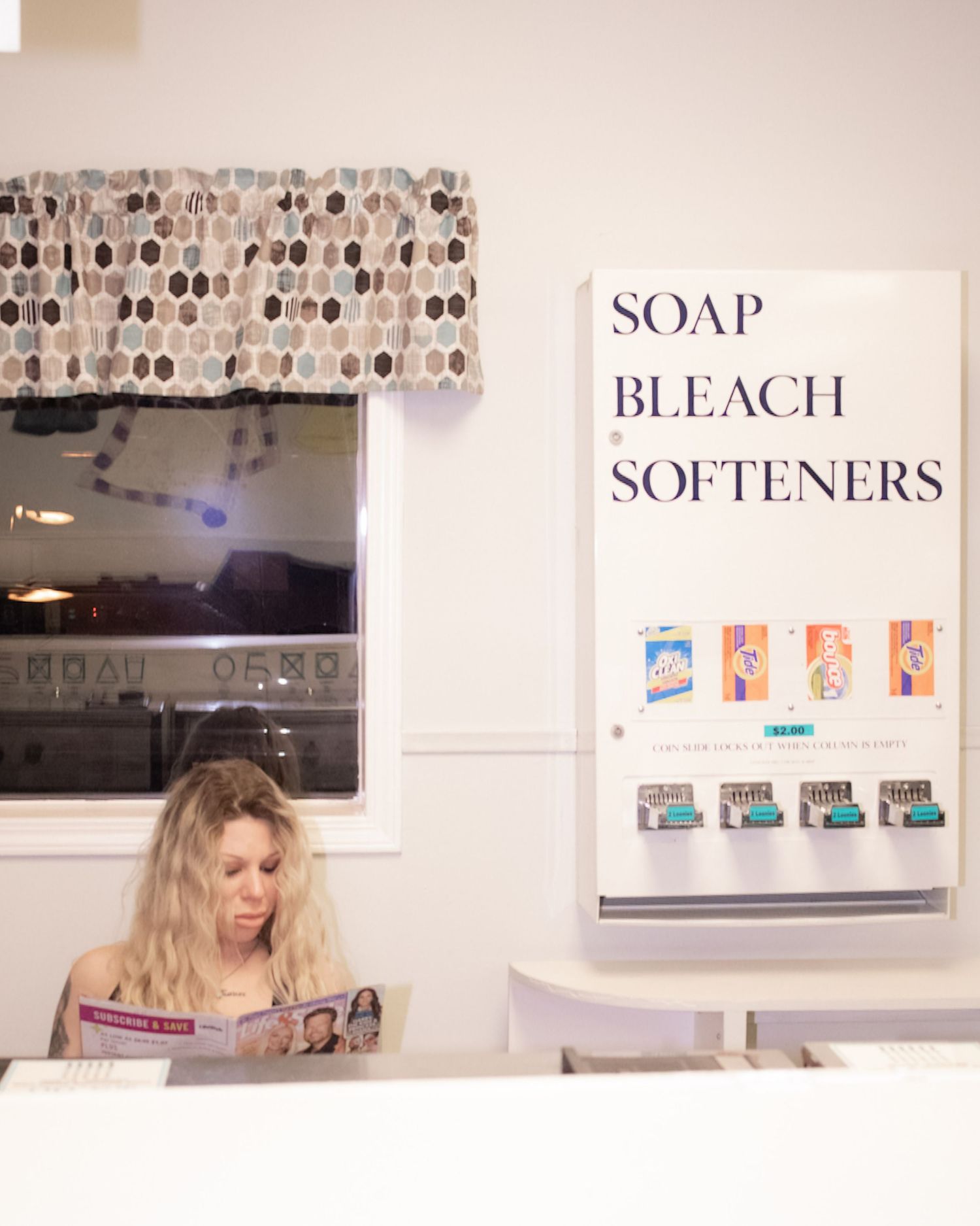 Blonde woman reading magazine at laundromat counter beneath soap bleach and softeners vending sign.