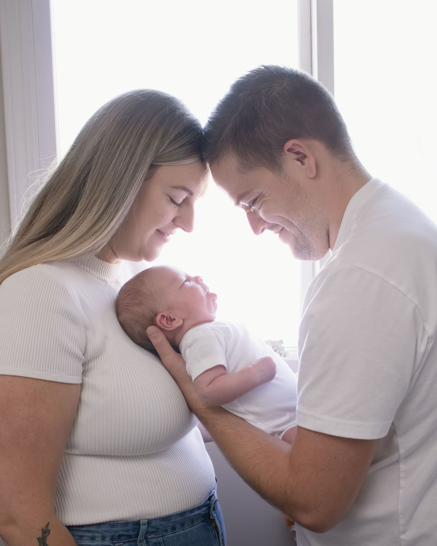 New parents tenderly hold their newborn baby together, touching foreheads, wearing white tops near a bright window.