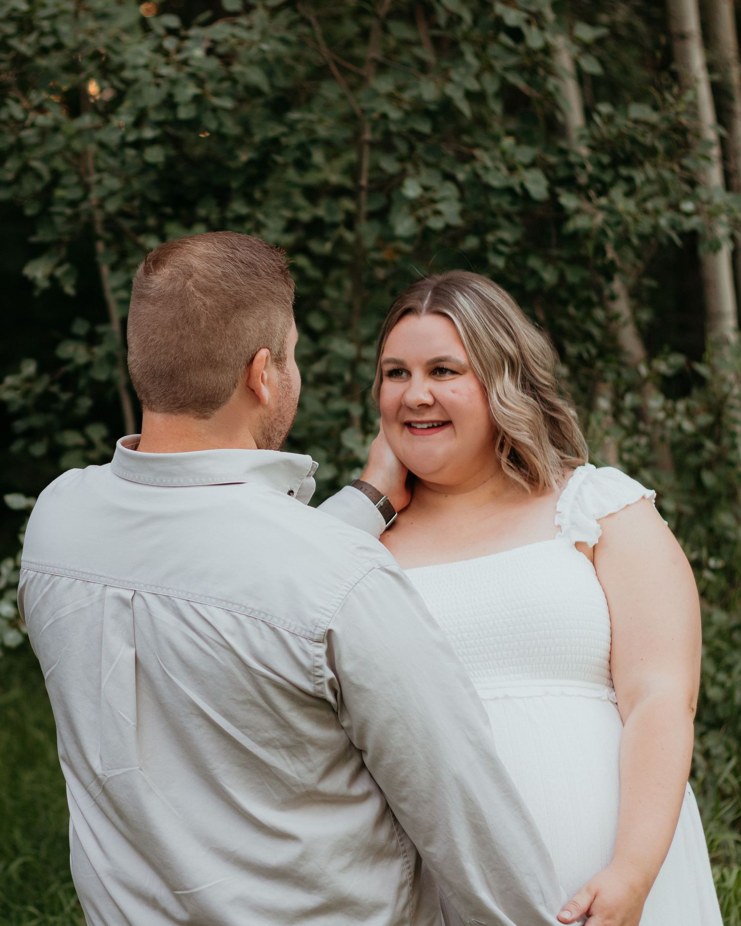 Smiling pregnant woman in white dress with partner touching her face outdoors.