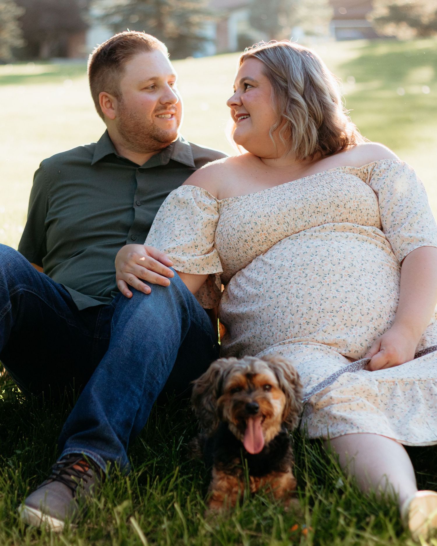 Smiling couple sitting on grass with their small dog during a golden hour maternity photoshoot.