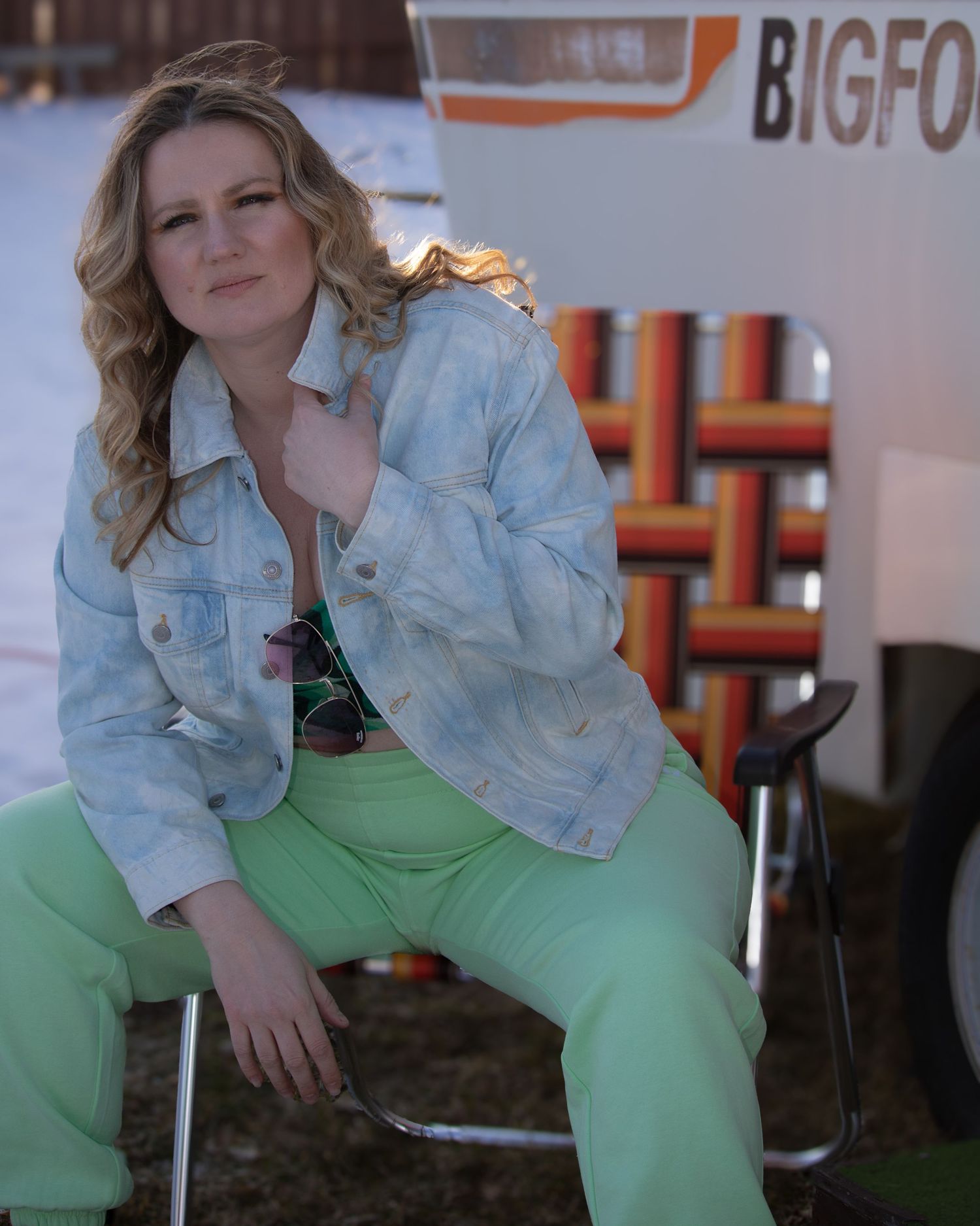 Blonde woman in mint green pants and tie-dye denim jacket sitting in front of a Bigfoot camper.