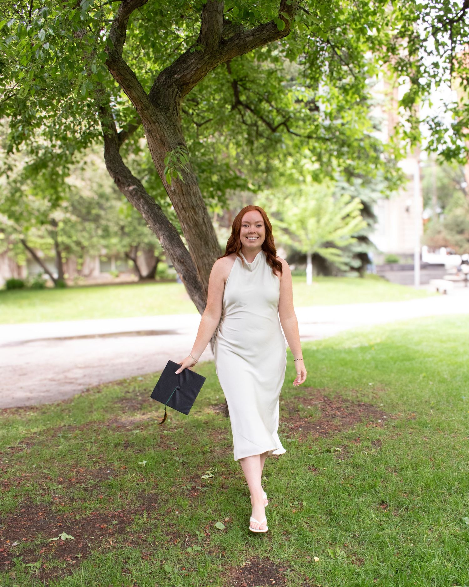 A smiling woman in a white midi dress holds a graduation cap while walking on green grass under a large tree.