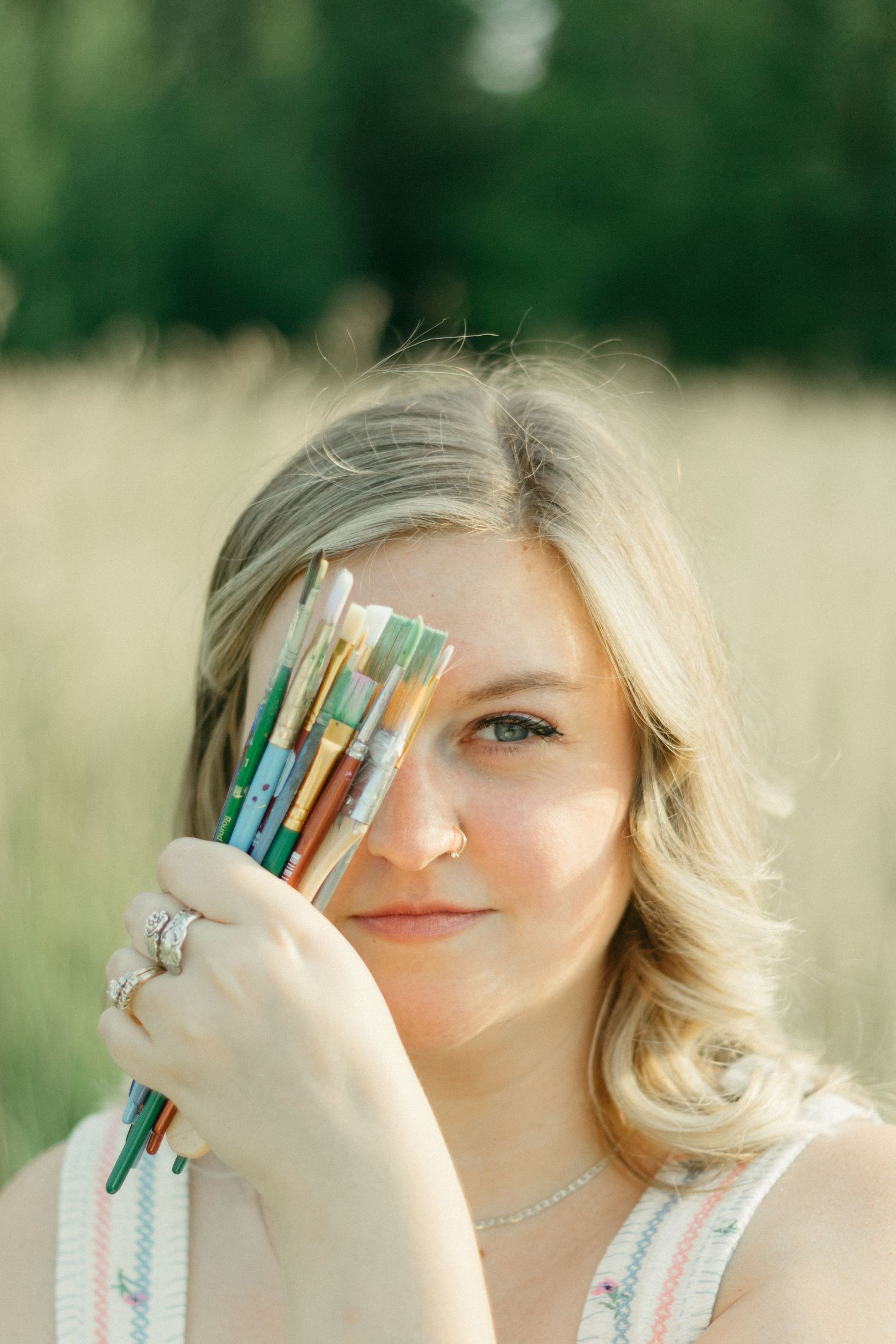 Blonde woman holding colorful paintbrushes over one eye in a sunlit field.