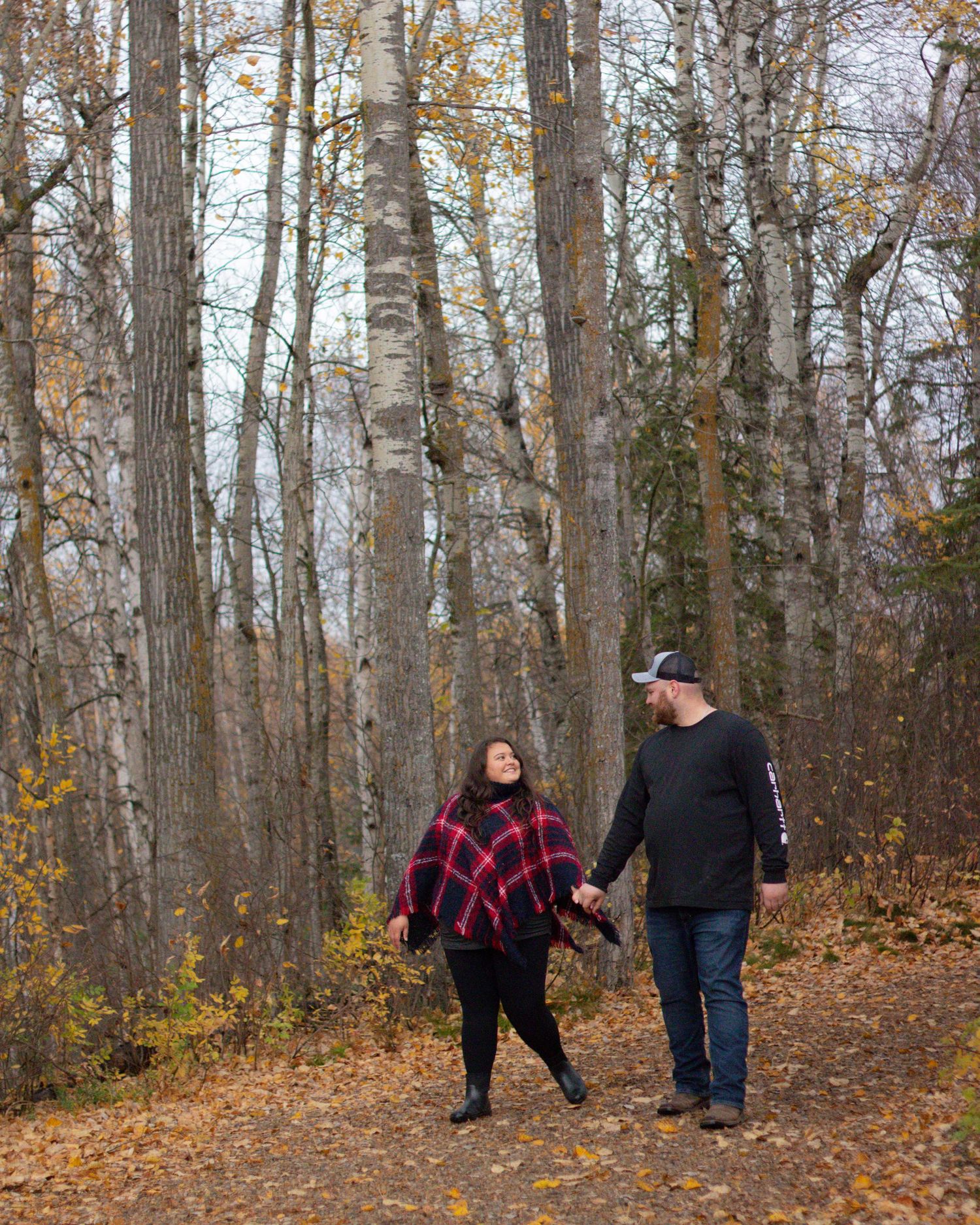Couple holding hands walking on a leaf-covered forest path surrounded by tall trees in autumn.