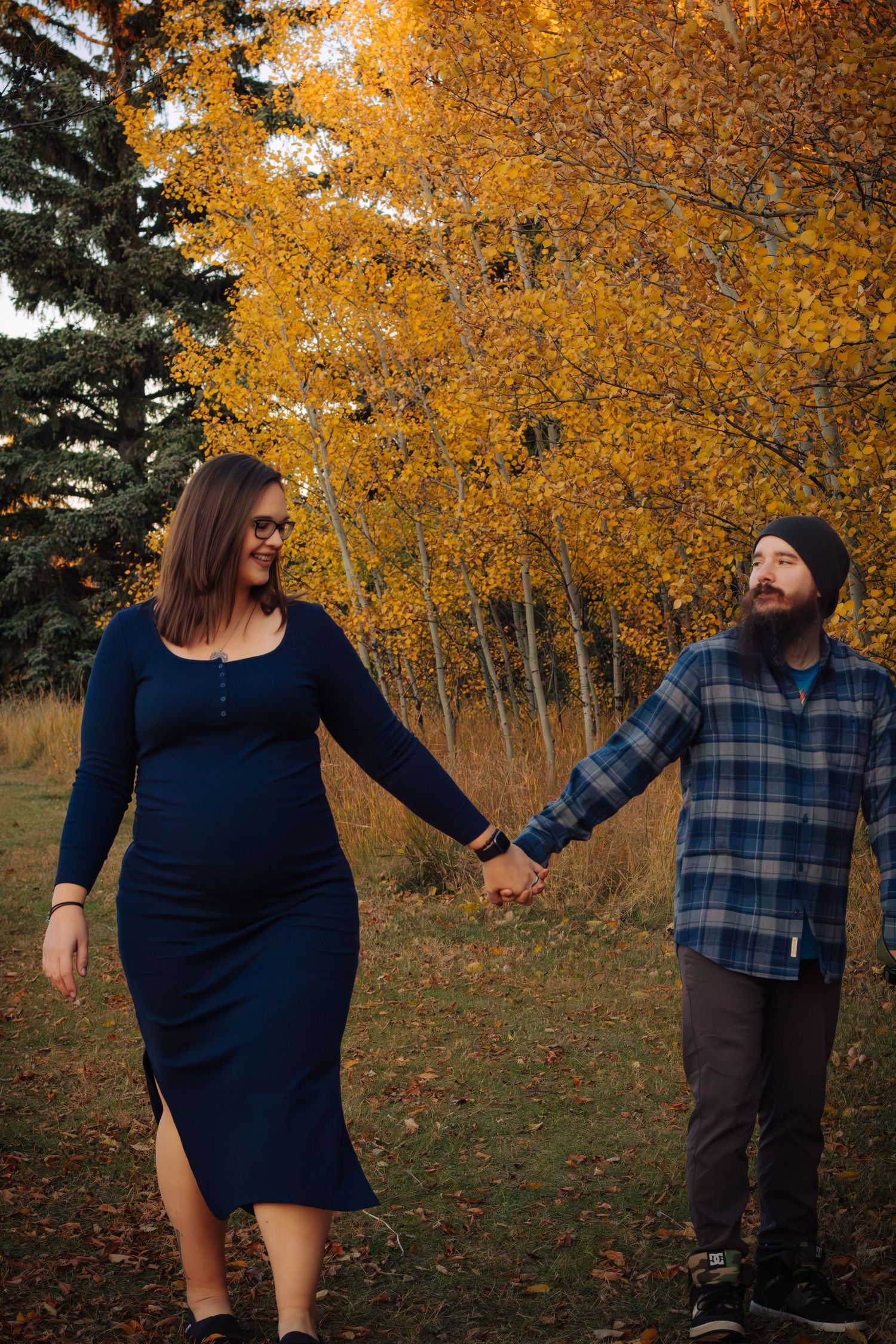 Pregnant couple holding hands walking together among vibrant yellow autumn trees.