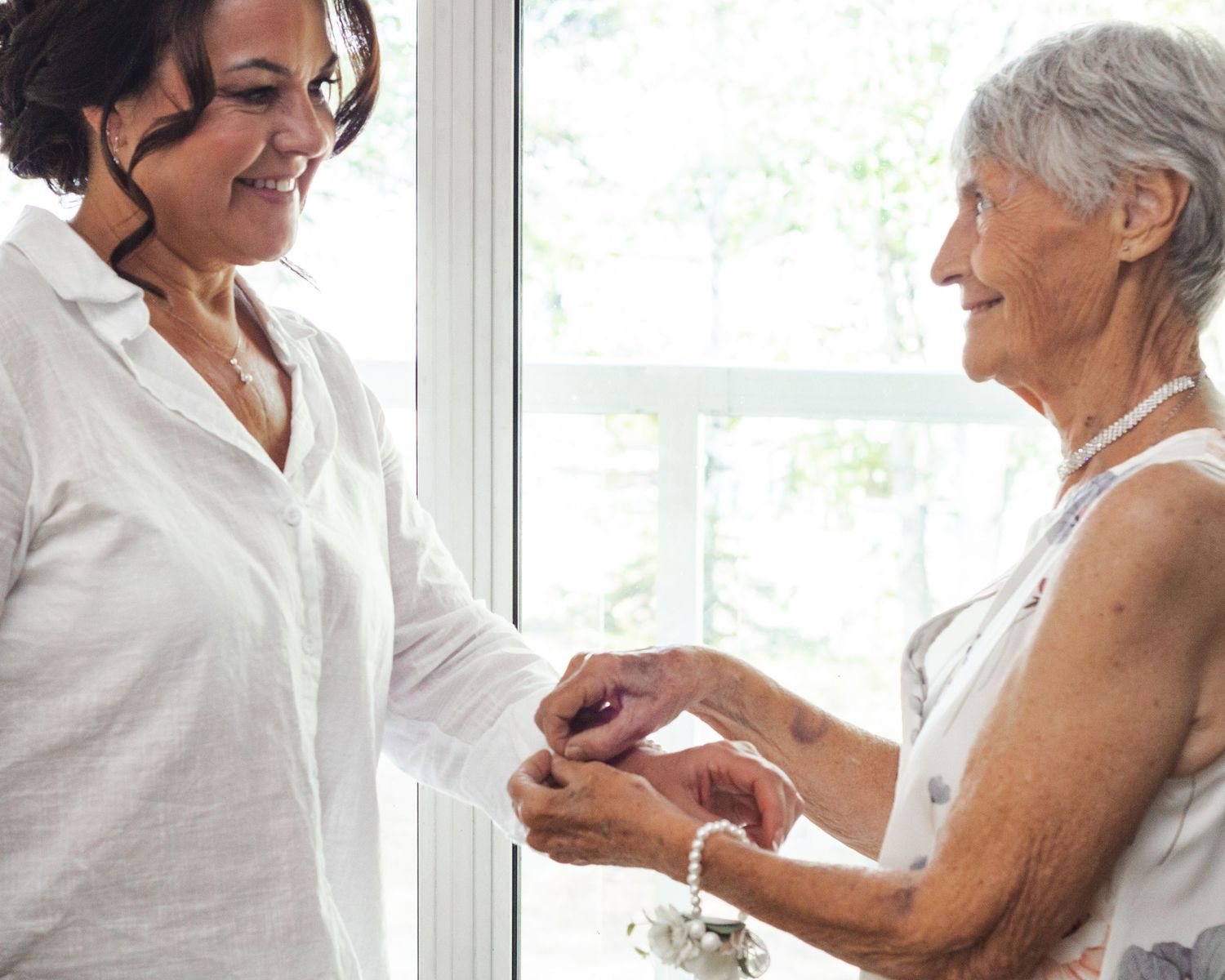 Two women smiling and sharing a tender moment by a bright window indoors.