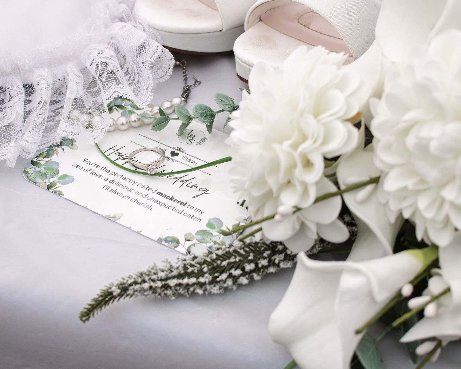 Elegant wedding flat lay featuring white bridal bouquet, heels, lace garter, and floral wedding card on white fabric.