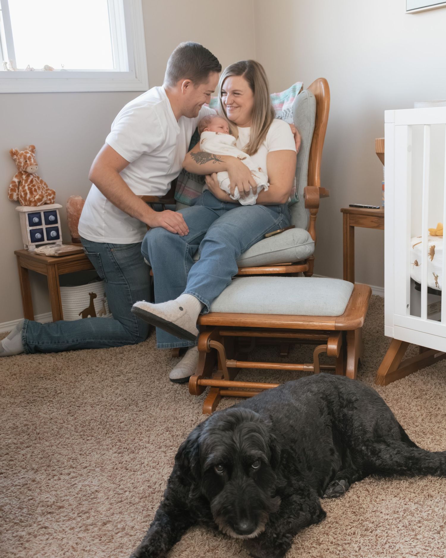 New parents admire their newborn in a nursery rocking chair while their black dog rests on the carpet nearby.