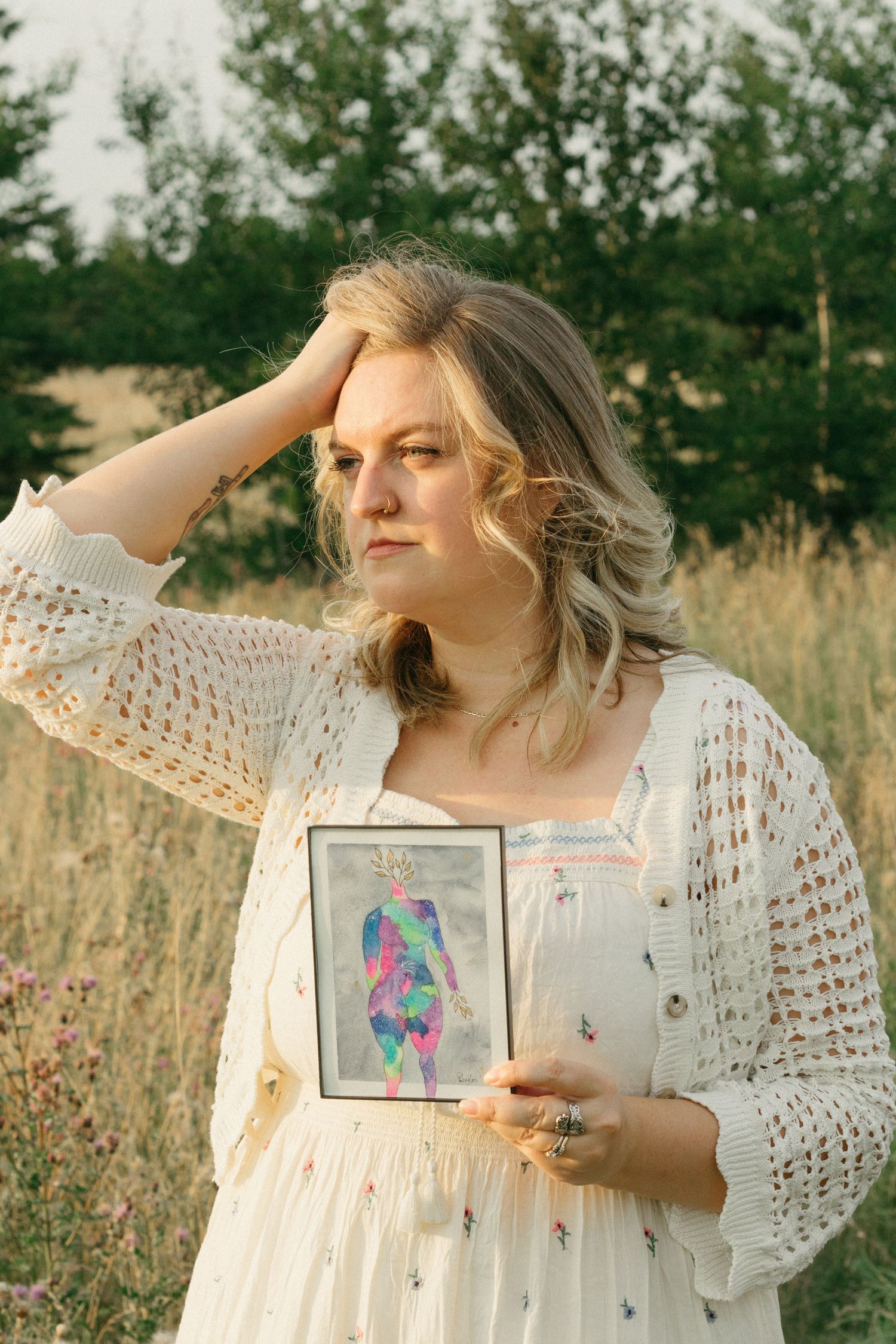 Woman in white crochet dress holding vibrant body-positive artwork in a sunny field.