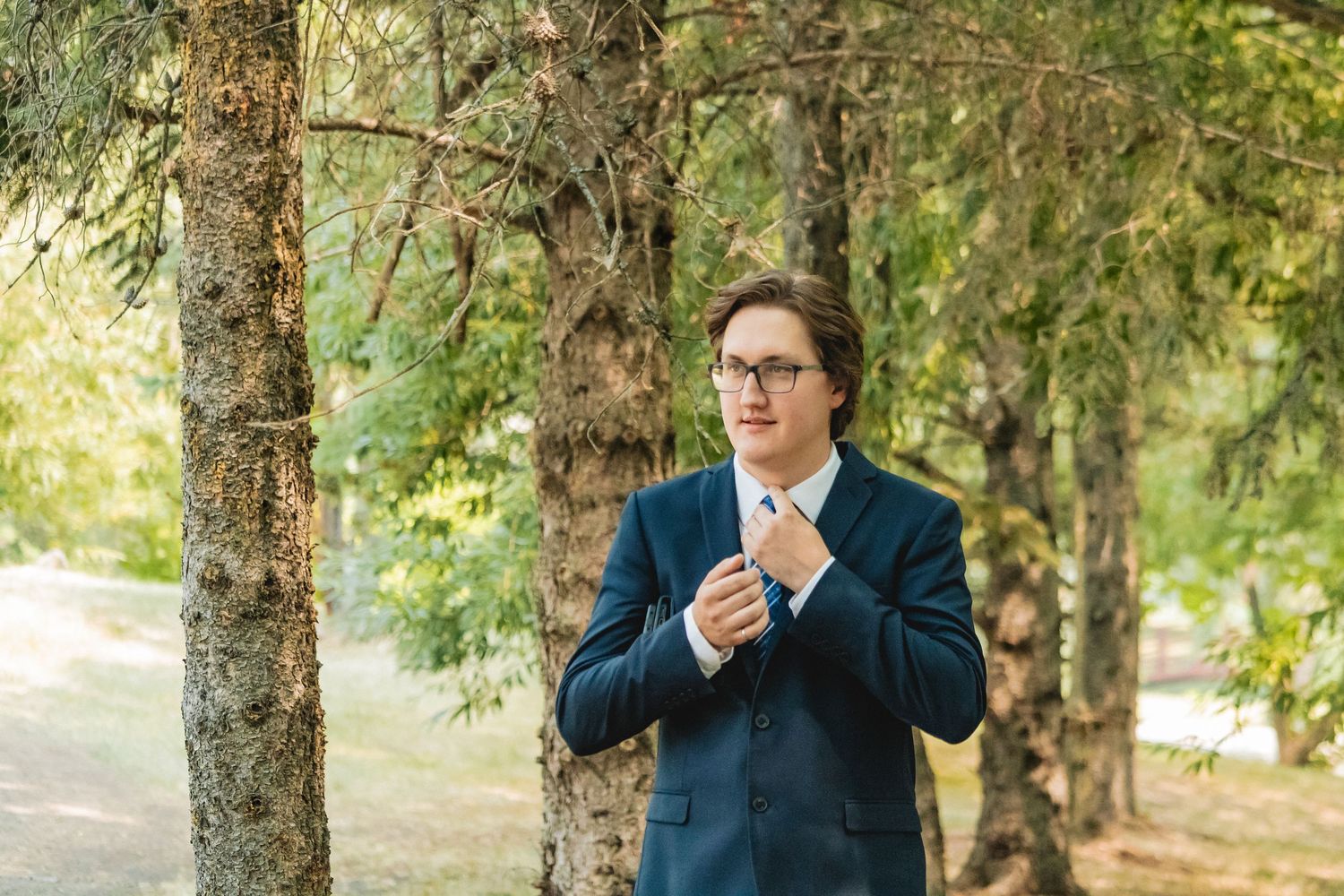 Groom in navy blue suit adjusting his tie among tall trees in an outdoor setting.