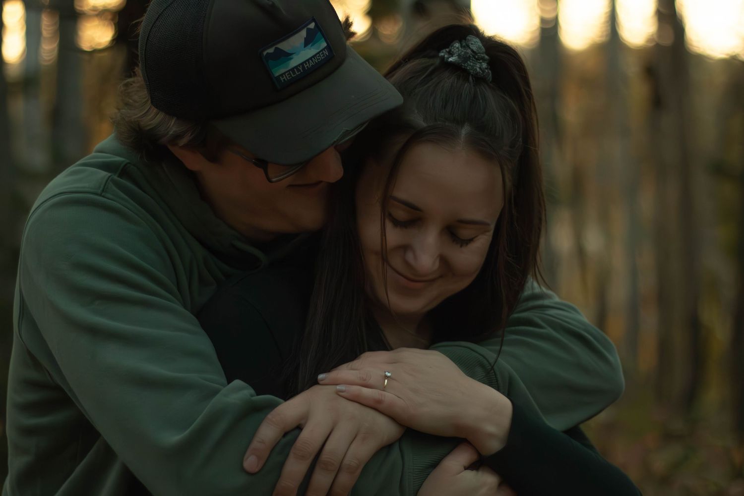 Couple embraces in autumn forest, woman smiling and showing engagement ring on her finger.