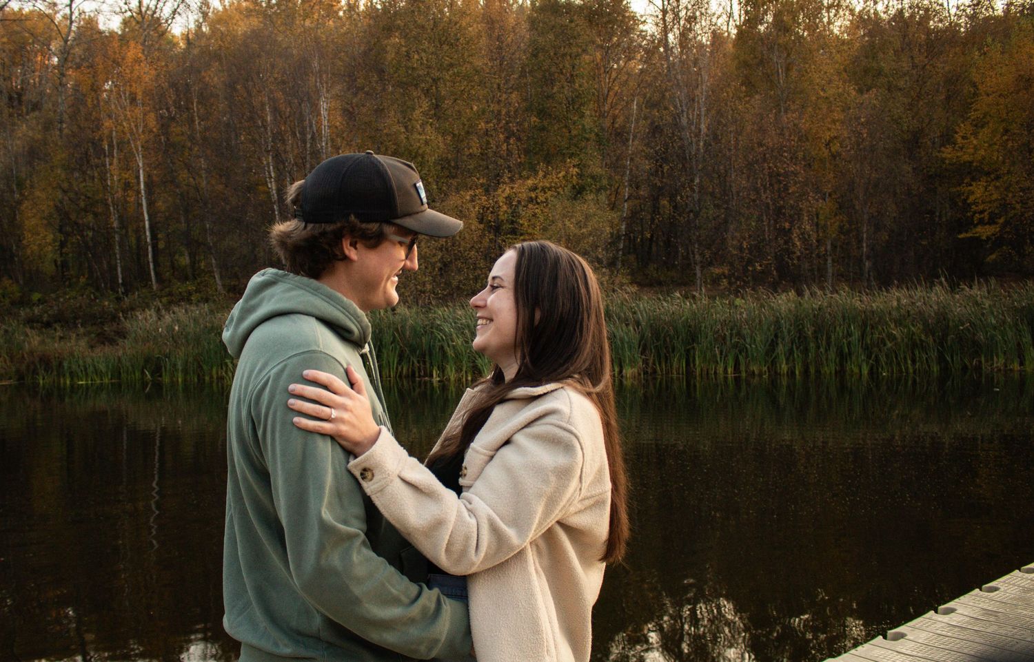 Couple smiling at each other on a dock by a lake surrounded by autumn foliage.