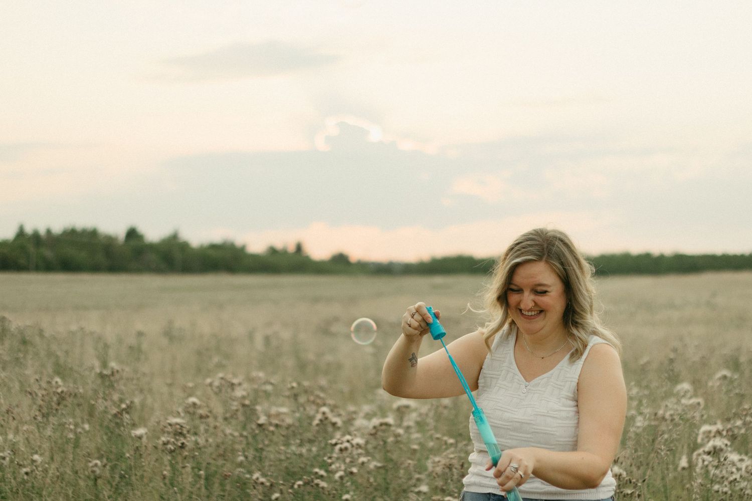 Happy woman blowing bubbles in a wildflower meadow at sunset, holding a blue bubble wand.