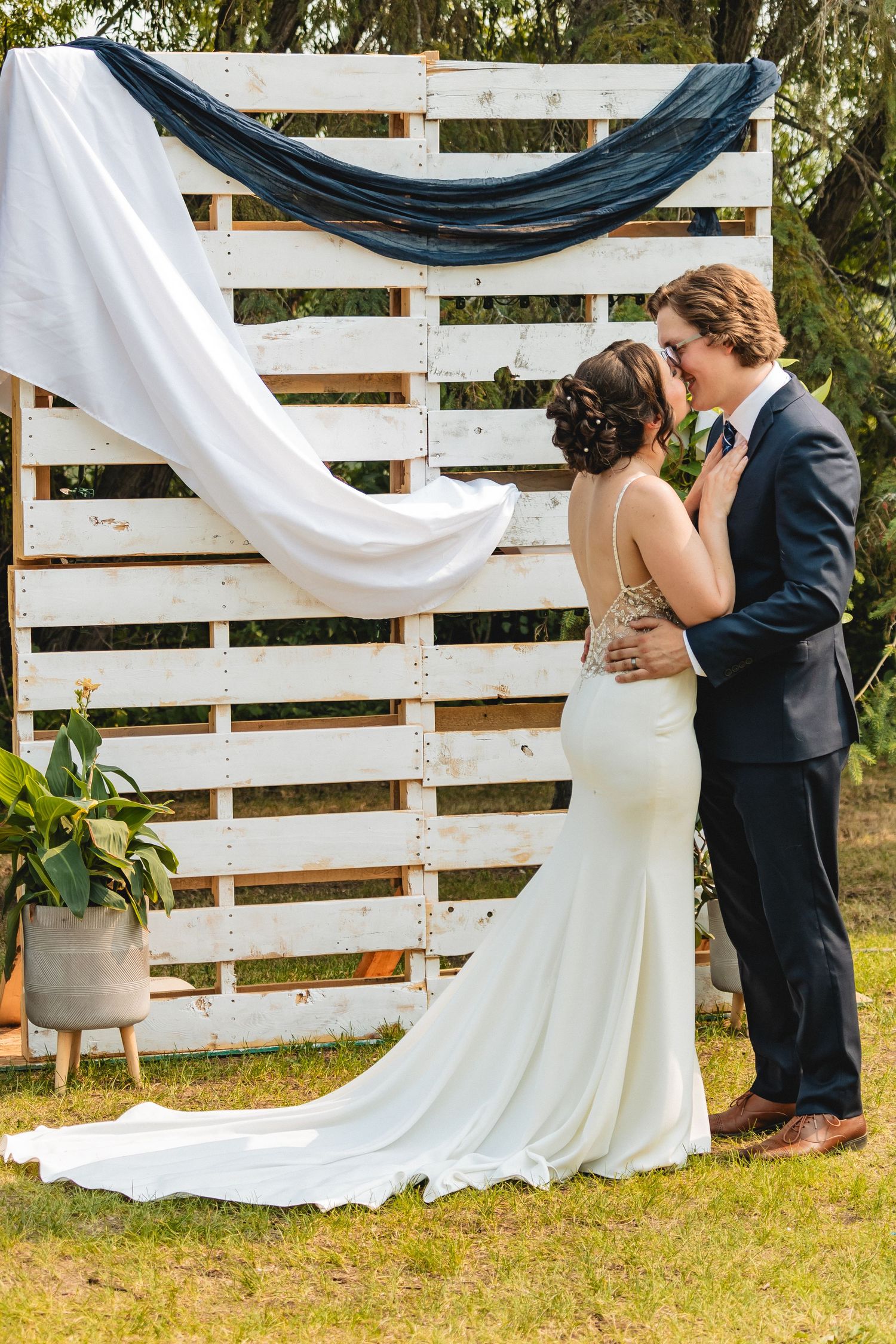 Bride and groom kissing at outdoor wedding altar decorated with white pallet backdrop and draped fabric.