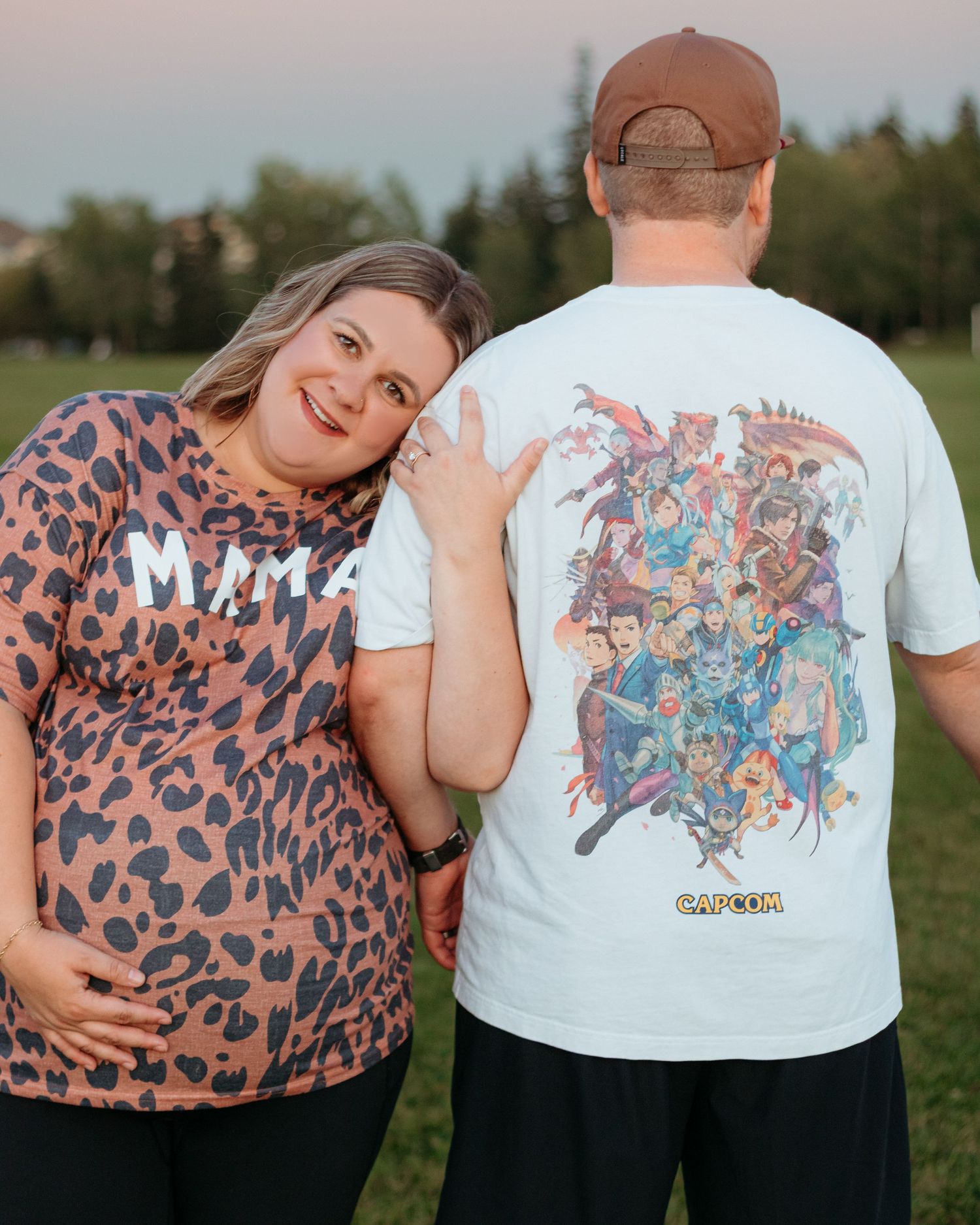 Smiling pregnant woman in leopard MAMA shirt posing with partner wearing Capcom graphic tee outdoors.