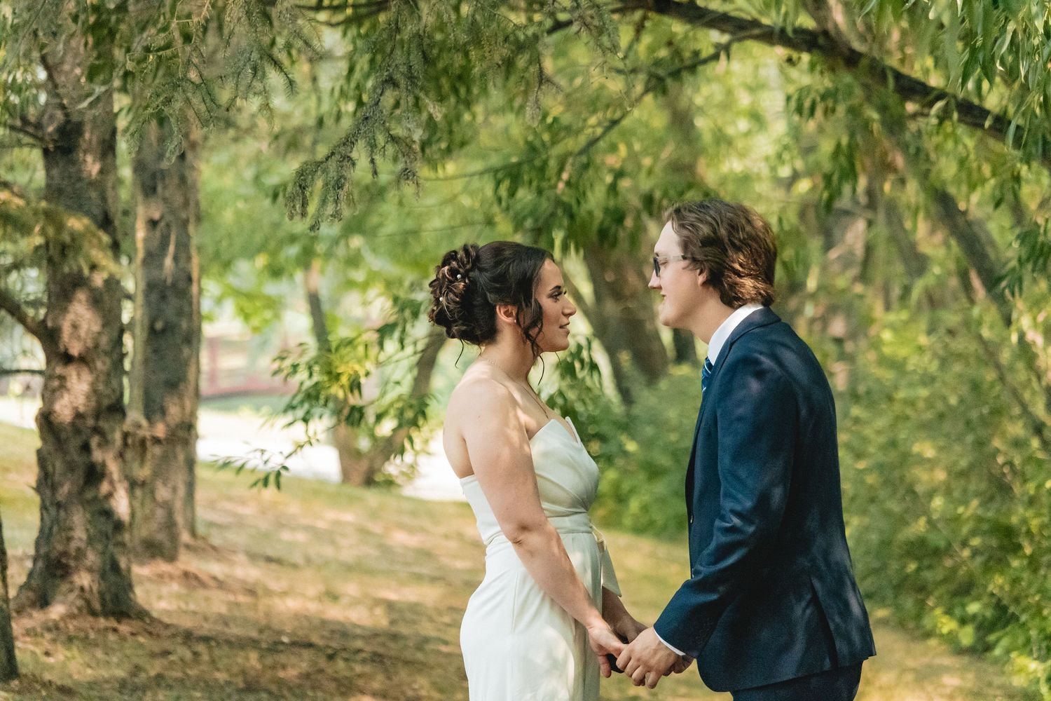 Bride and groom holding hands and smiling at each other under lush green trees on their wedding day.