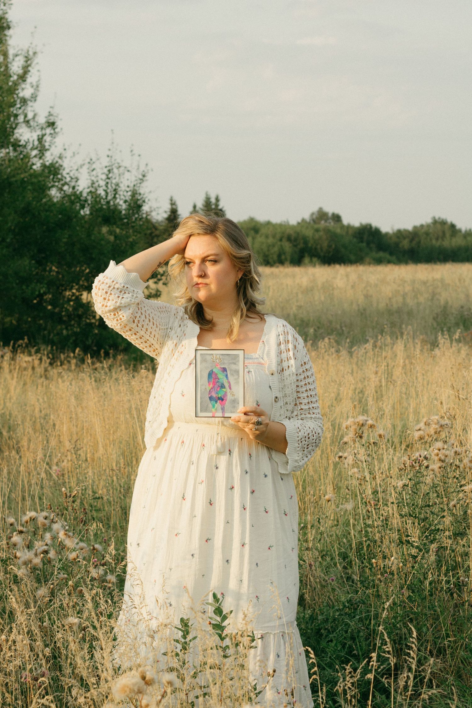 Blonde woman in white boho dress holding framed colorful art in a wildflower field.