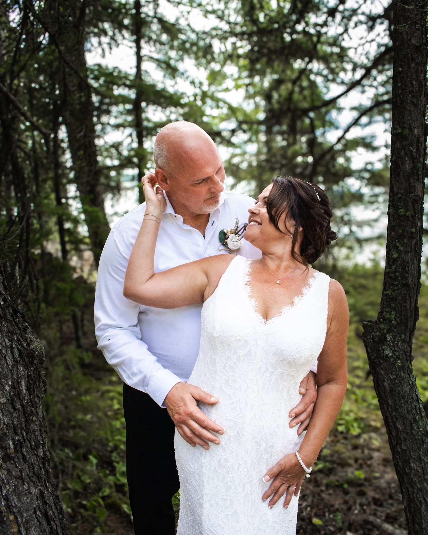 Bride and groom share a romantic moment among pine trees, groom tenderly touching bride's face.