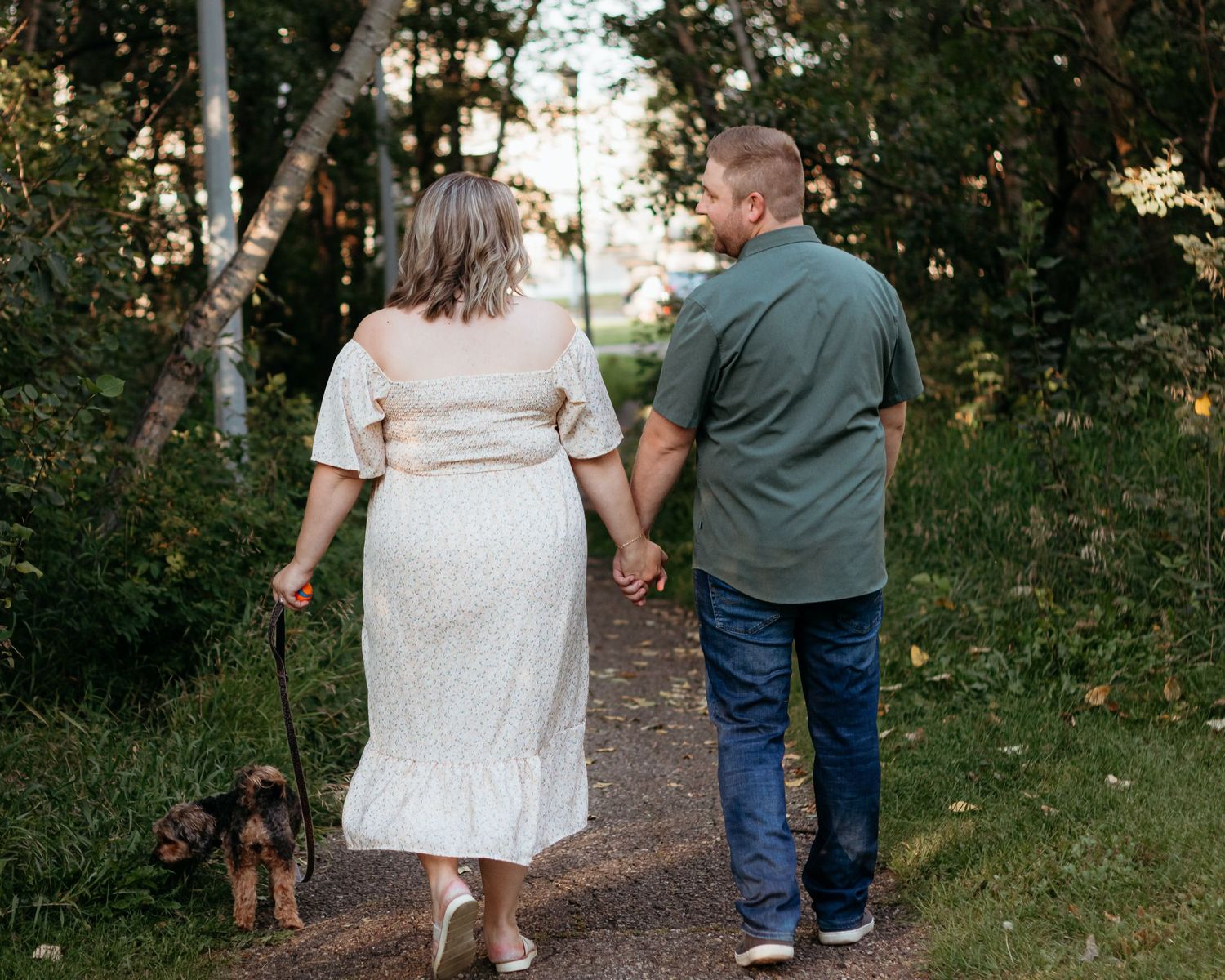 Couple walking a small dog on a path through lush green trees, holding hands.