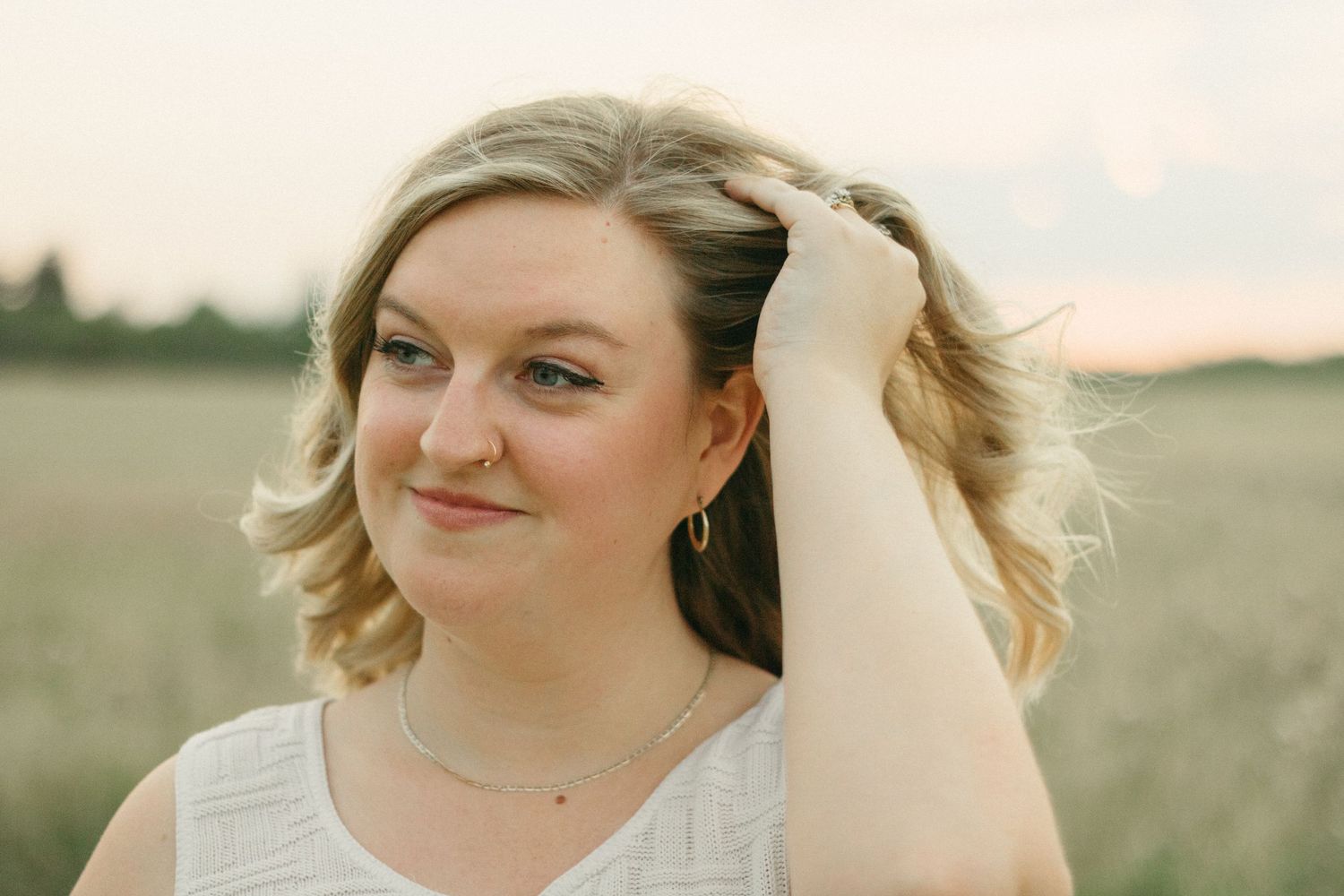 Close-up of a blonde woman with hoop earrings touching her hair in a golden field at sunset.
