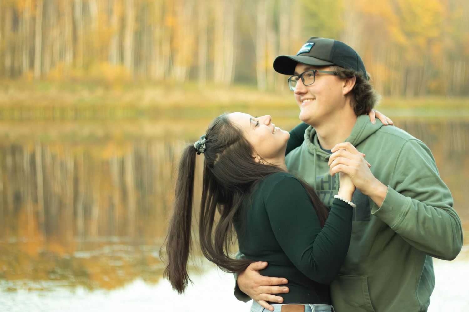 Happy couple dancing and laughing together by a lake with vibrant fall foliage behind them.