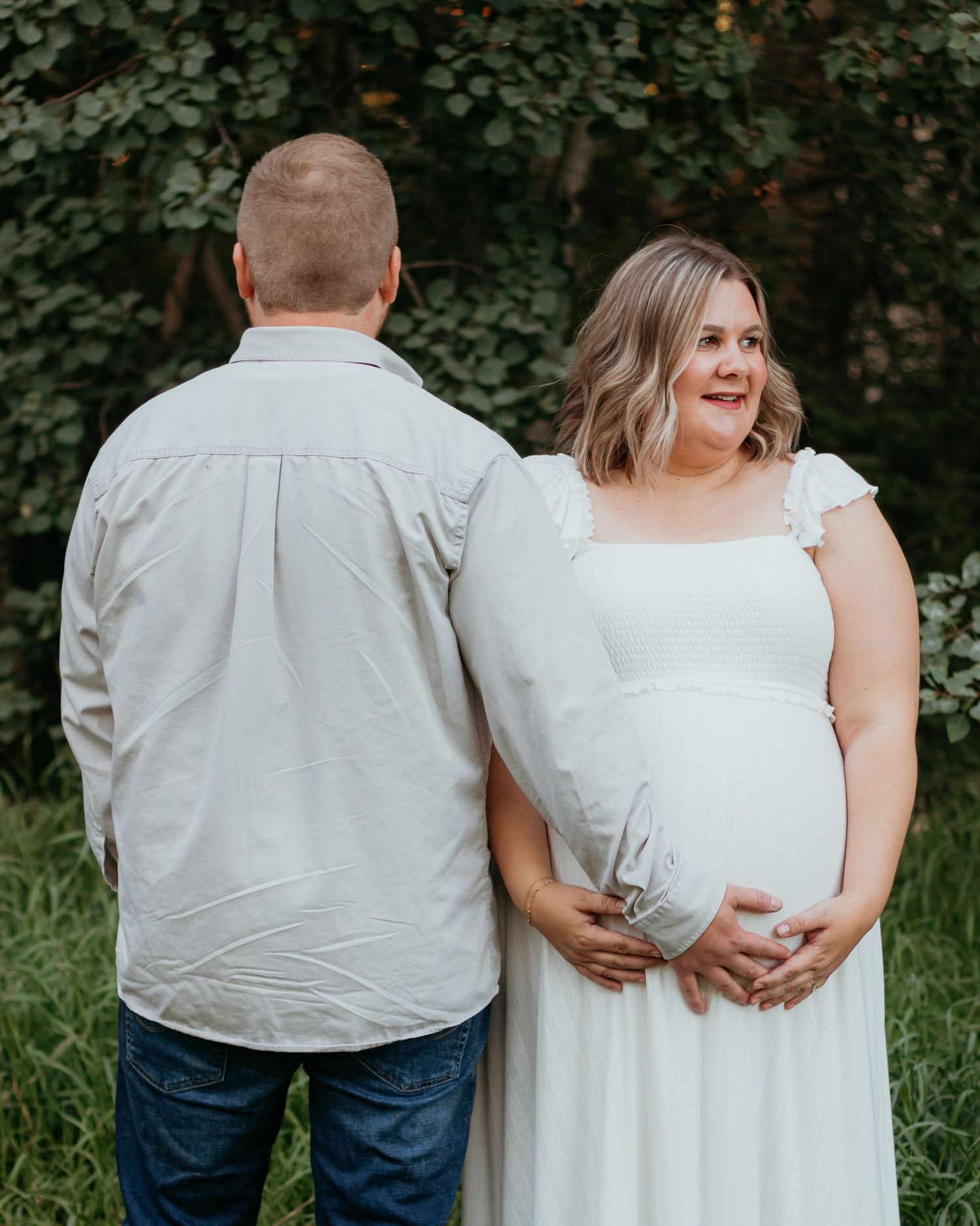 Pregnant woman in white gown holding baby bump while standing with partner in front of green ivy.