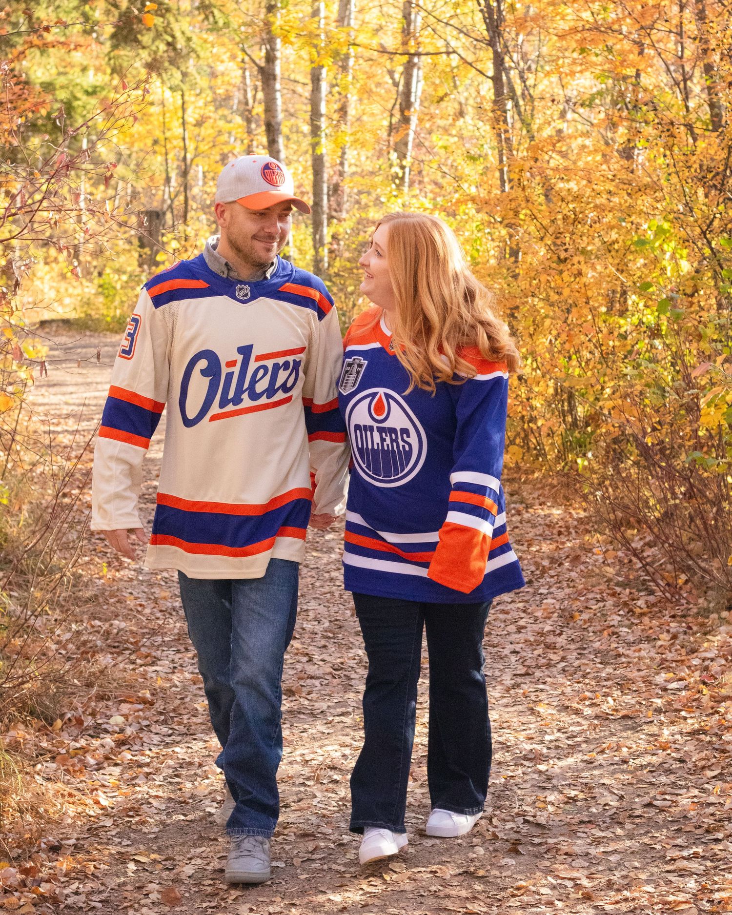 Couple walking hand in hand on autumn trail wearing Edmonton Oilers hockey jerseys among golden fall foliage.