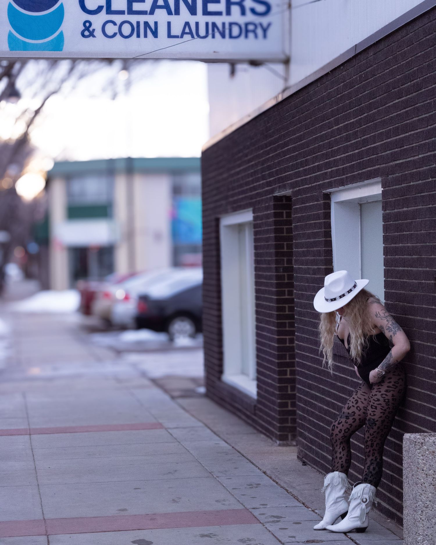 Blonde woman in leopard print outfit and white hat leaning against brick wall outside coin laundry.