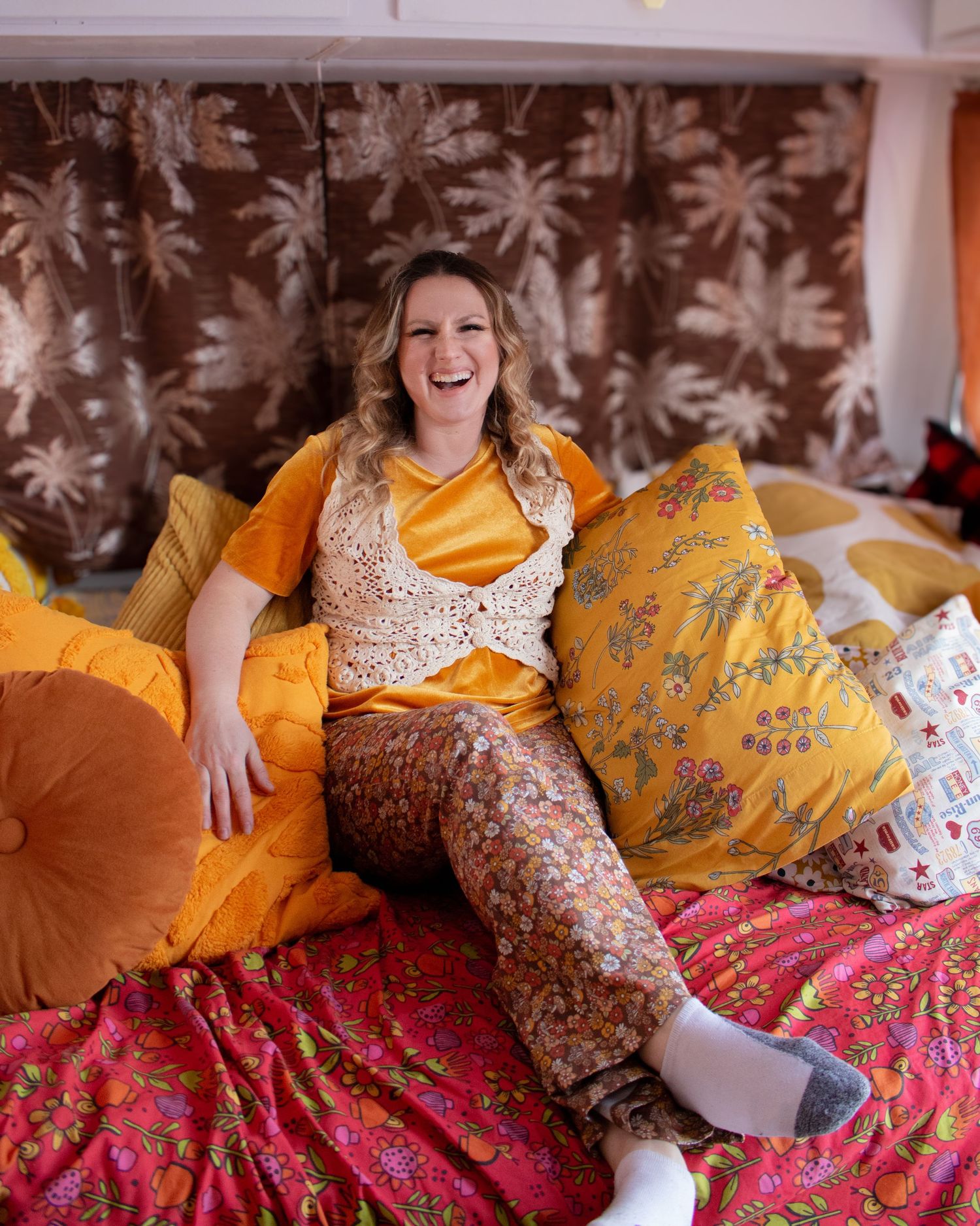 Laughing woman in yellow satin top and floral pants sitting among colorful pillows in a retro camper interior.