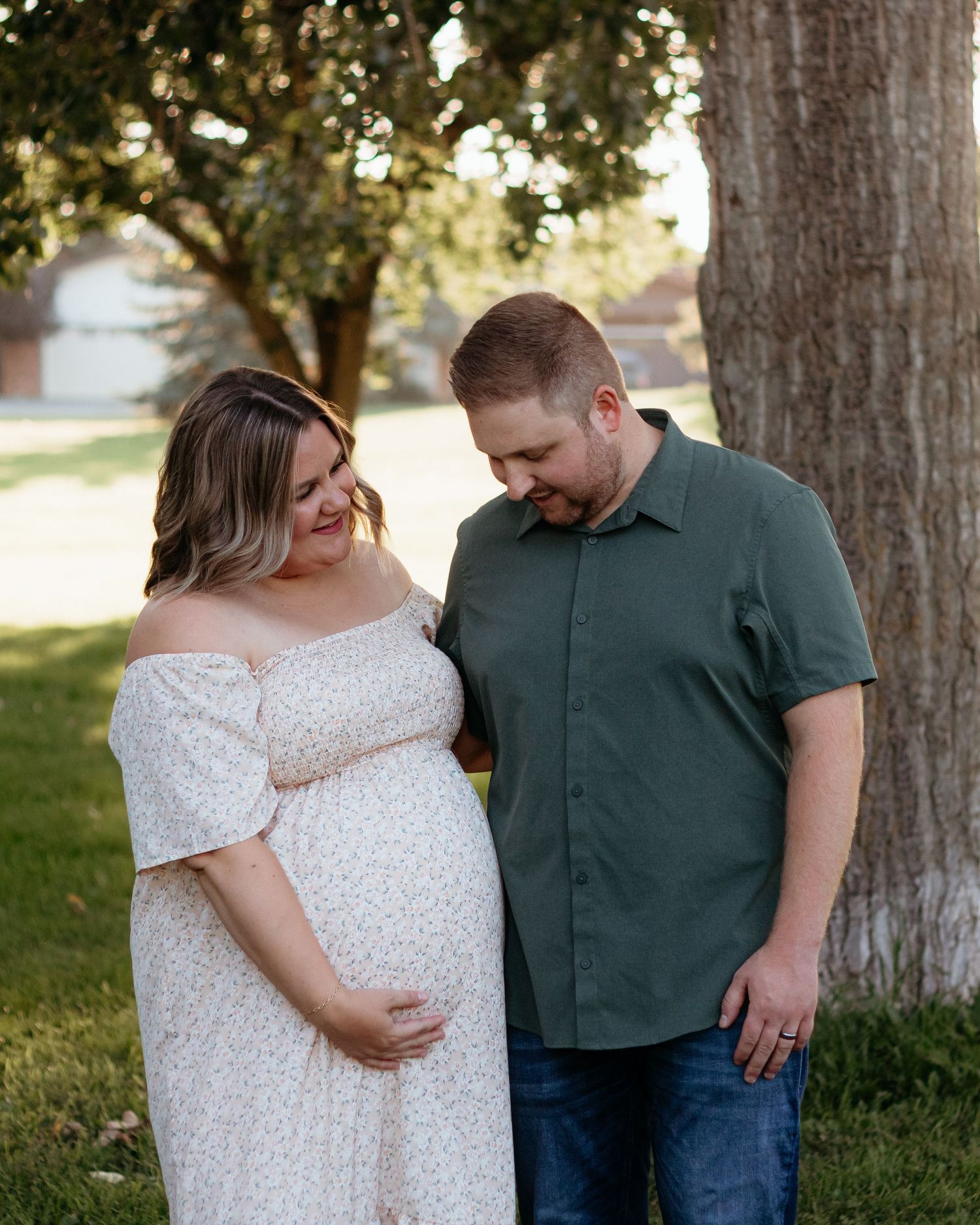 Expectant couple smiling at baby bump under a large tree during golden hour maternity photoshoot.