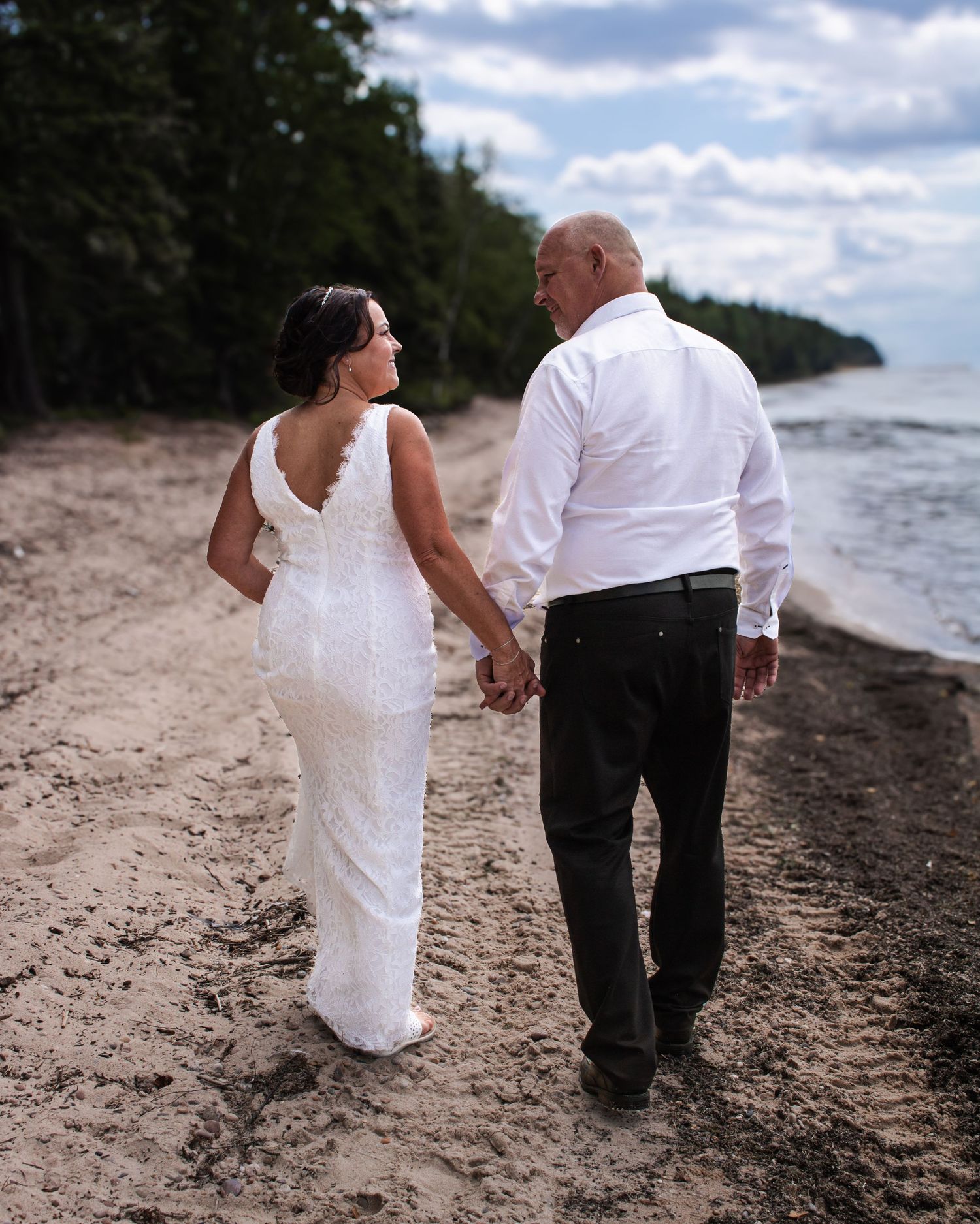 Bride and groom walking hand-in-hand along a sandy beach, bride in white lace gown, groom in white shirt.