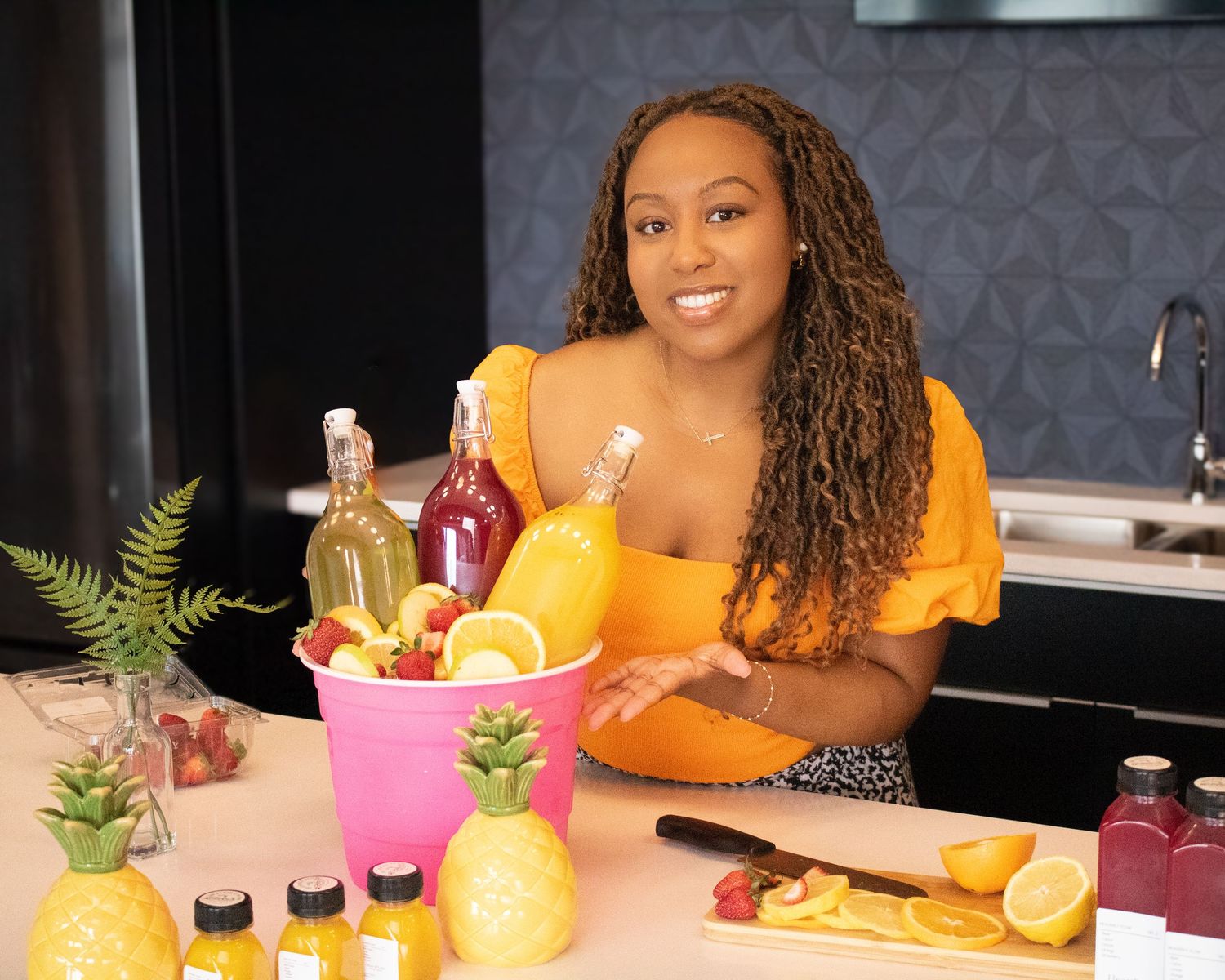 Smiling woman in yellow top poses with tropical fruit display and colorful bottles in modern kitchen.