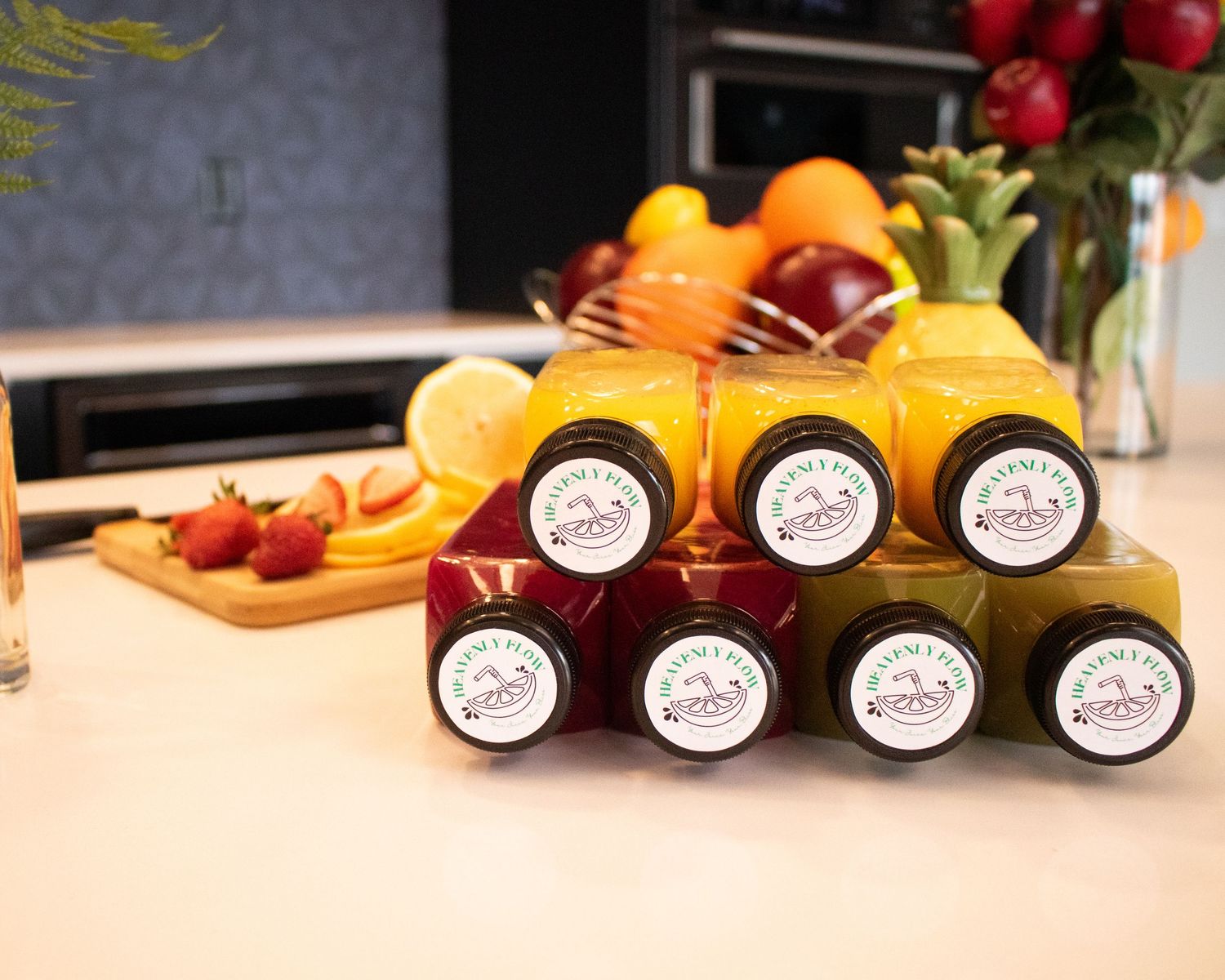 Six bottled fresh juices arranged on counter with tropical fruits and berries in background.
