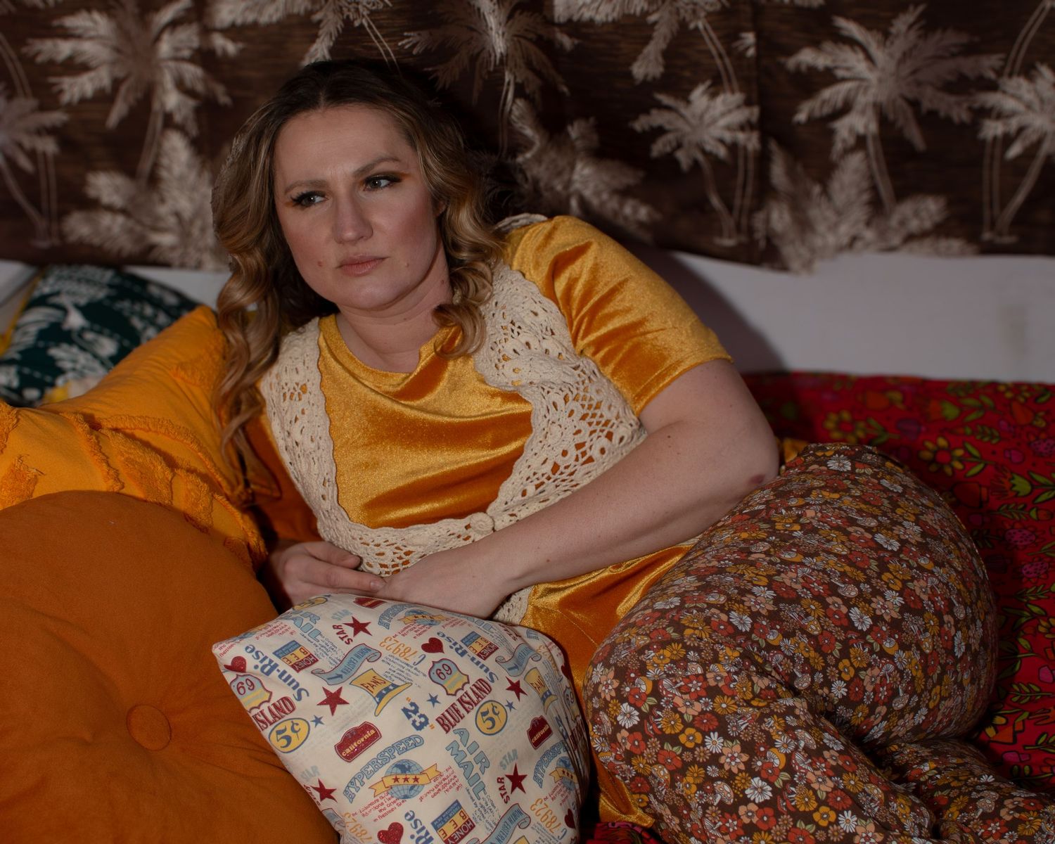 Woman in gold velvet dress reclining on colorful floral bedding inside vintage camper interior.