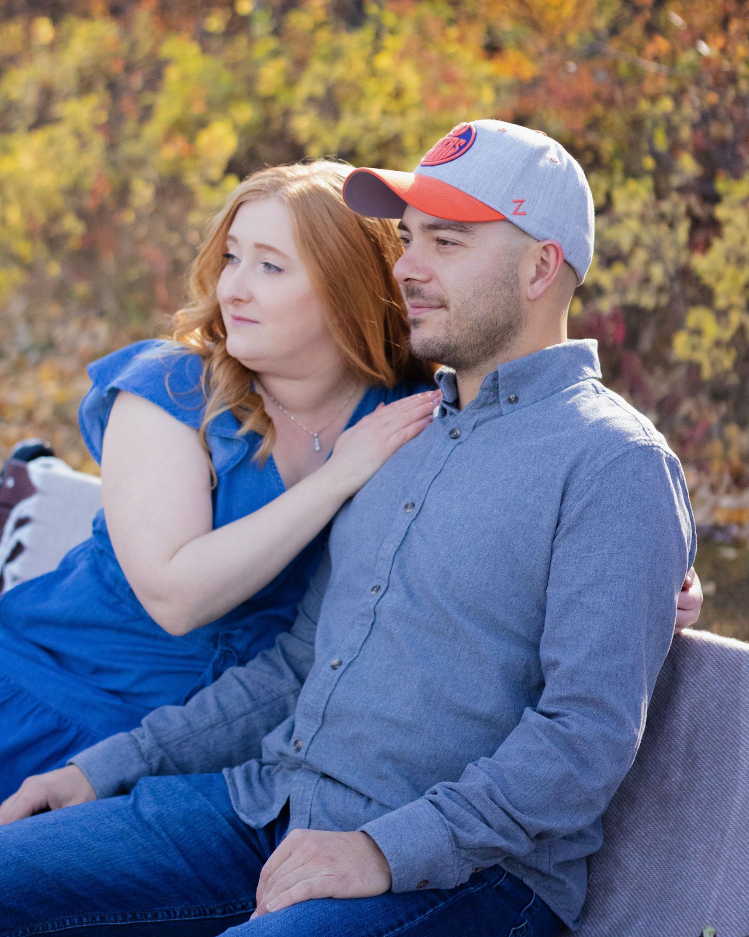 Couple sitting together outdoors with colorful fall foliage, woman in blue dress resting hand on mans shoulder.